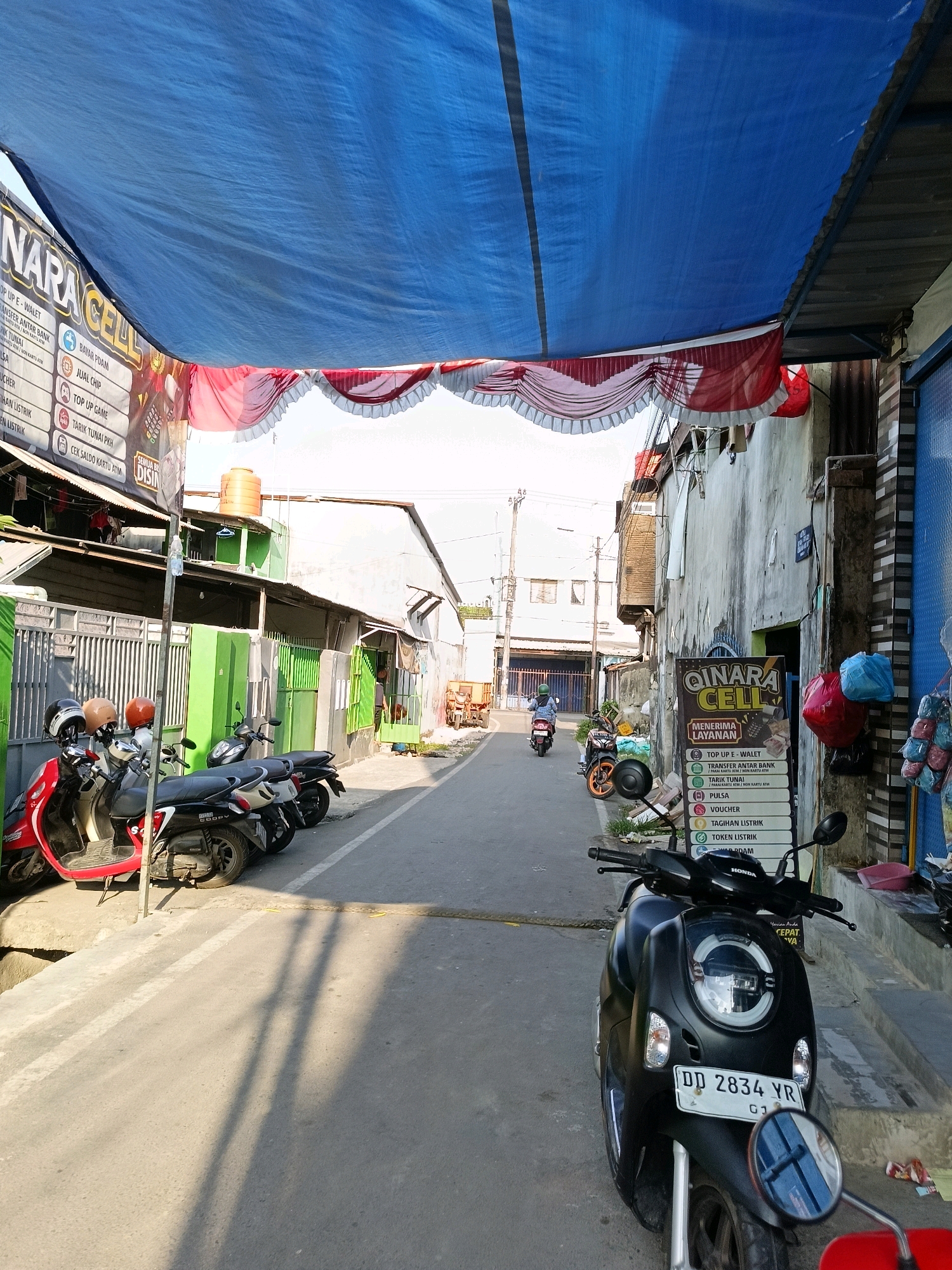 The image depicts a street scene in Makassar, Indonesia, likely in a commercial area. A blue tarp is stretched overhead, providing shade, with red and white decorative fabric draped below it. On the left side of the street, several scooters are parked. A sign for "QINARA CELL" is prominently displayed on the right, listing various services such as mobile top-ups, money transfers, bill payments, and voucher sales. The buildings flanking the street appear to be shops or small businesses. Further down the street, a few individuals on motorcycles are visible, suggesting ongoing activity. The environment appears to be a typical urban street with a mix of commercial establishments and residential structures. The time of day appears to be daytime, with sunlight casting shadows on the road.