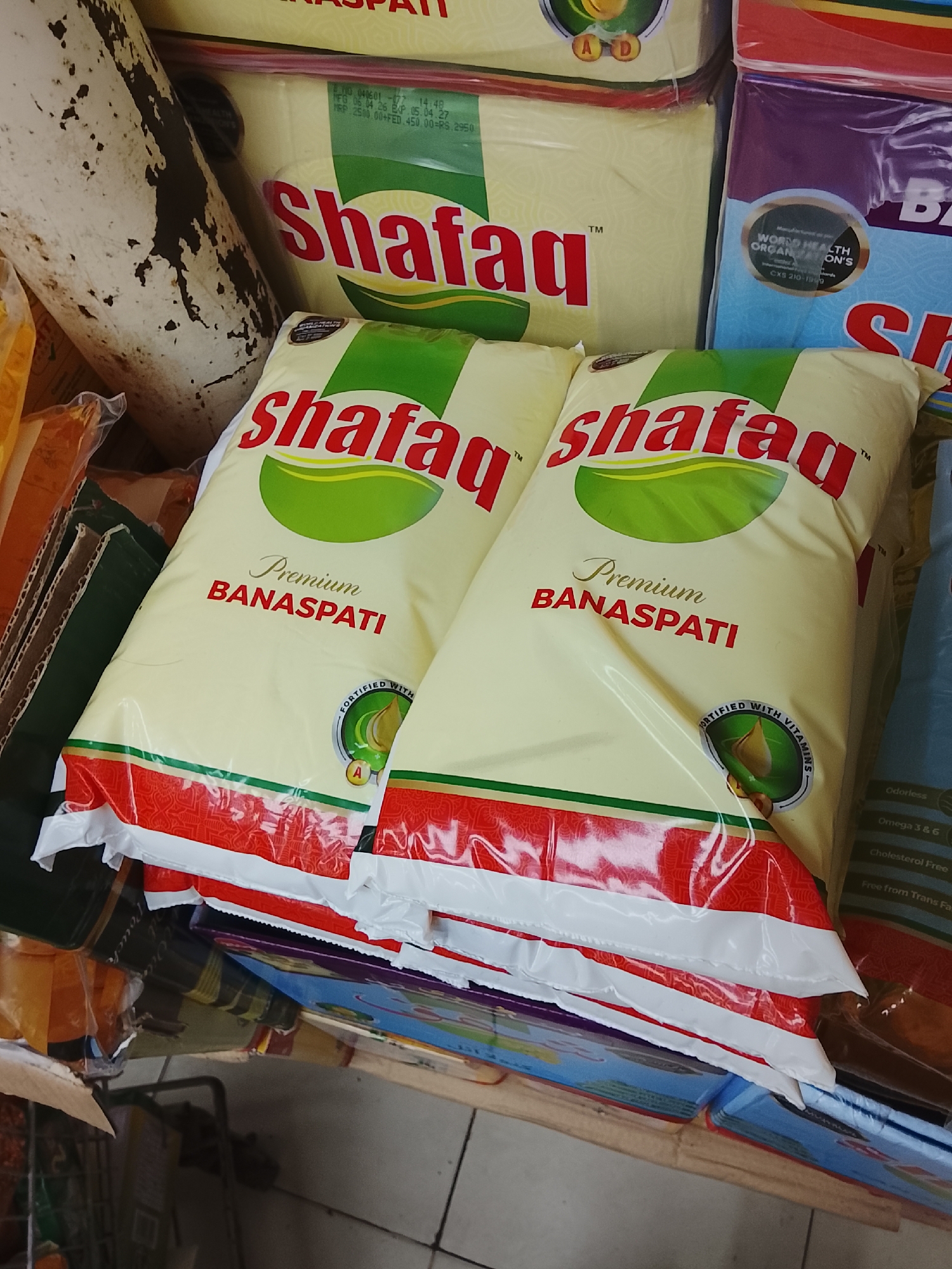The image shows several packages of "Shafaq Premium Banaspati" cooking oil stacked on a shelf, likely in a shop or market. The packages are mostly white with red trim and feature the "Shafaq" brand name in red letters above a green leaf design. Text on the packages indicates that the product is fortified with vitamins and has a premium quality. A package of "Shafaq" cooking oil is above the two packages in the foreground. Visible on the other packages is the text, "WORLD HEALTH ORGANIZATION'S," indicating that these oils meet WHO standards for food safety. The scene appears to be indoors, potentially in a retail setting. The picture was taken in the city of Gulgasht Colony, Pakistan.