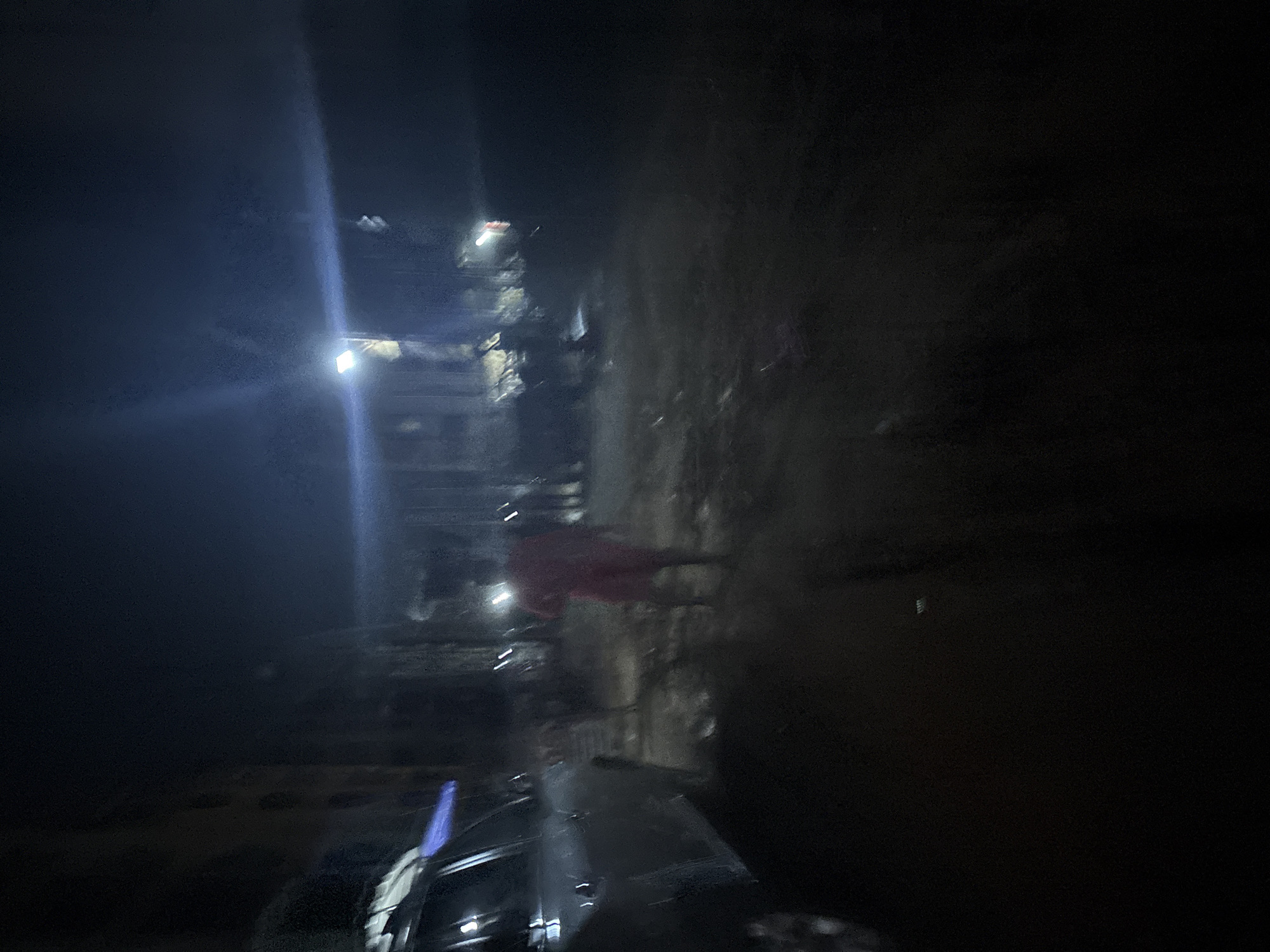A nighttime street scene in Onitsha, Nigeria, is depicted with limited visibility due to darkness and artificial lighting. In the foreground, the left side of a silver SUV is partially visible, with its headlights on, casting a faint glow. A person dressed in red shorts and a lighter-colored top is walking away from the camera in the middle ground, illuminated by a bright overhead light source. Behind this individual, other figures and structures are dimly visible, suggesting an urban environment. The ground appears to be dirt or gravel, with some debris scattered around. The overall impression is one of a dimly lit, possibly outdoor, gathering or street activity at night.