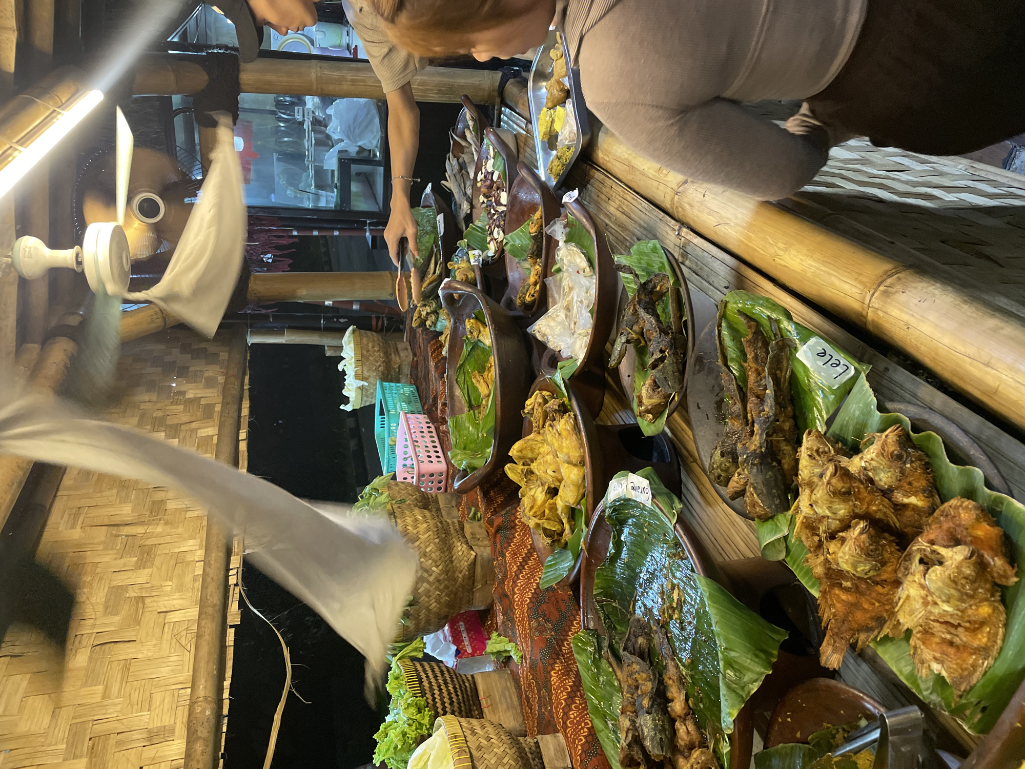 The image captures a food stall in North Jakarta, Indonesia. A variety of fried fish dishes are displayed in a row of earthenware and metal platters lined with banana leaves, accompanied by handwritten labels. The food stall is set up against a wall of woven bamboo. Two people are present, with one person reaching out to select food from the display. A fan spins in the foreground, adding to the outdoor market atmosphere.