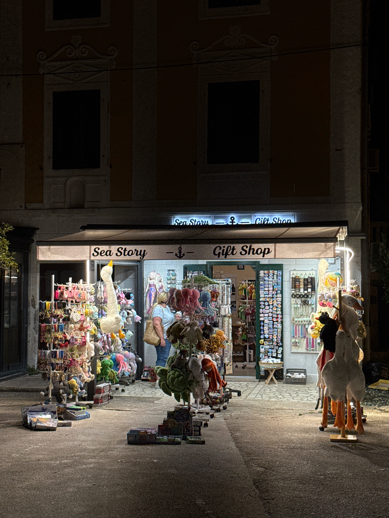 This image depicts a nighttime scene in front of a gift shop called "Sea Story - Gift Shop" located in the city of Novigrad, Croatia. The shop's awning is lit, illuminating a display of various merchandise. In the foreground, on the left side of the street, are racks filled with colorful plush toys, keychains, and other trinkets. On the right side, a display features large stuffed animals, including several geese with orange legs.
A woman, wearing a blue top and jeans, stands inside the shop, browsing the merchandise. She is carrying a straw bag over her shoulder. Inside the shop, more items are visible, including shelves of small boxes and what appear to be decorative items. The building itself has traditional architecture with dark windows and ornate detailing above the shop entrance. The street in front of the shop is paved with cobblestones and asphalt. The overall atmosphere is that of a small, charming tourist shop at night.