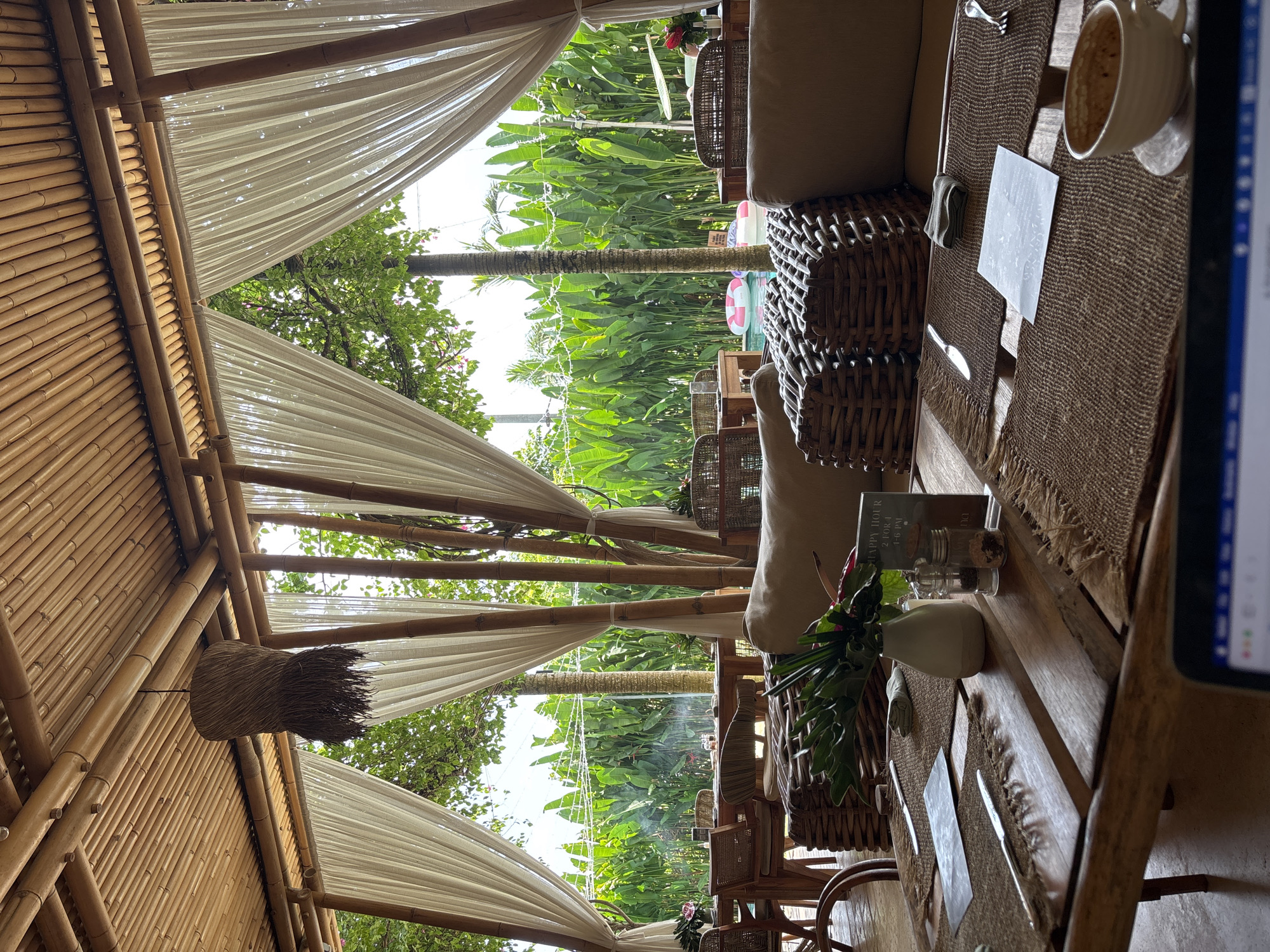 The image shows a partially outdoor seating area in what appears to be a restaurant or lounge. The location is in Kuta Selatan, Indonesia, evident from the tropical vegetation (banana trees, palm trees) visible in the background.
The seating area is covered by a bamboo frame structure draped with white fabric, providing shade and a semi-enclosed space. A decorative woven basket hangs from the bamboo structure.
The interior is furnished with wicker chairs and cushioned seating. A long wooden table is set with placemats, cutlery, small vases, and a cup of coffee. A book titled "HAPPY HOUR 2-FOR-1 4-6 PM" is visible on the table. A laptop computer is partially visible in the foreground.
A pink and white inflatable ring can be seen through the background vegetation, likely near a pool. No people are visible in the frame.