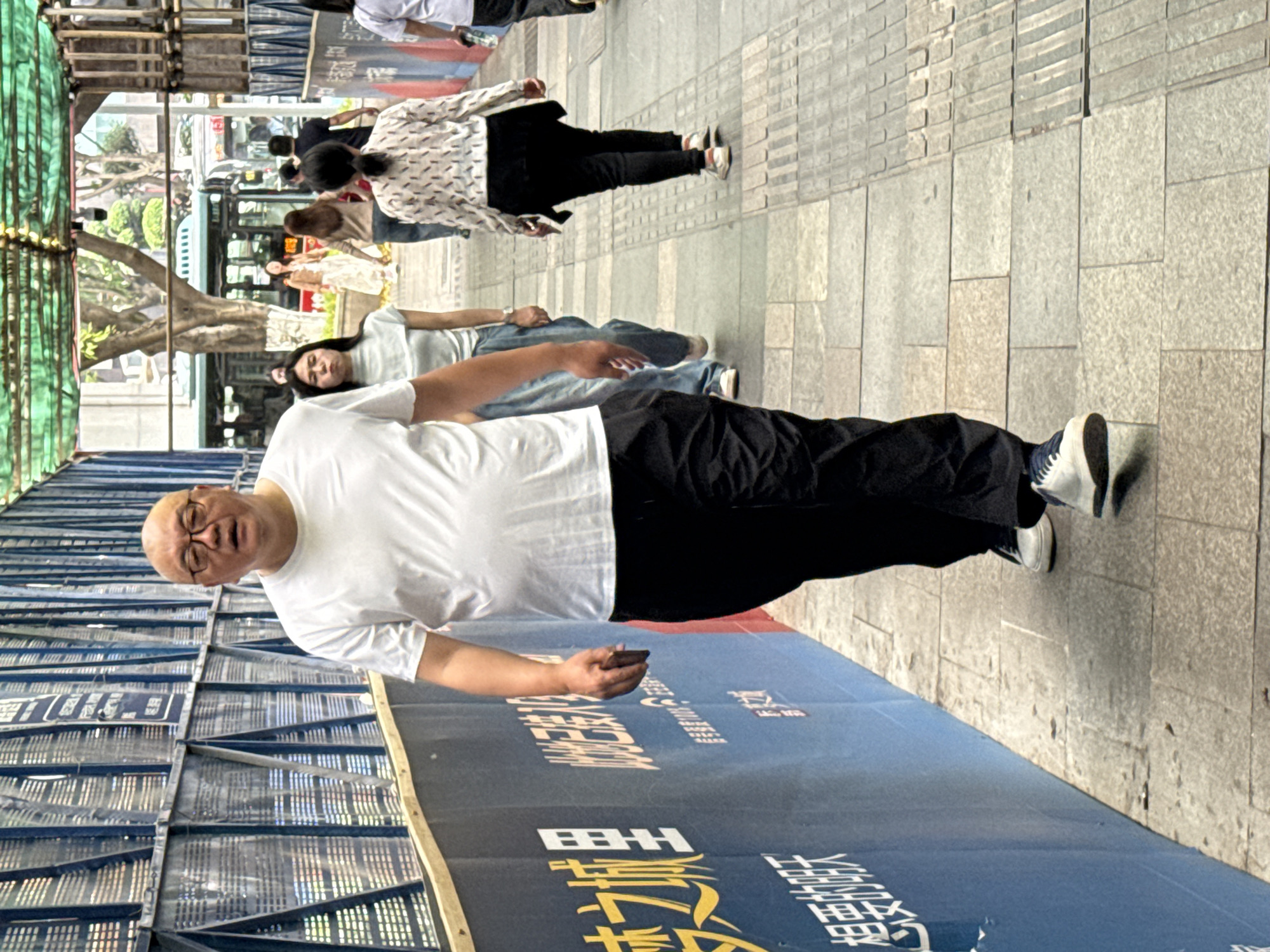 The image shows a man wearing a white t-shirt, black pants, and glasses, standing on a sidewalk next to a blue construction barrier with Chinese writing. In his hand, he holds a dark-colored object, possibly a phone. Behind him, other people walk along the sidewalk, including at least two women in light-colored shirts and jeans. In the background, there are trees and a bus, suggesting an urban setting in 龙溪街道, China. Construction scaffolding is visible on the left side of the frame. The sidewalk is paved with rectangular stone tiles.