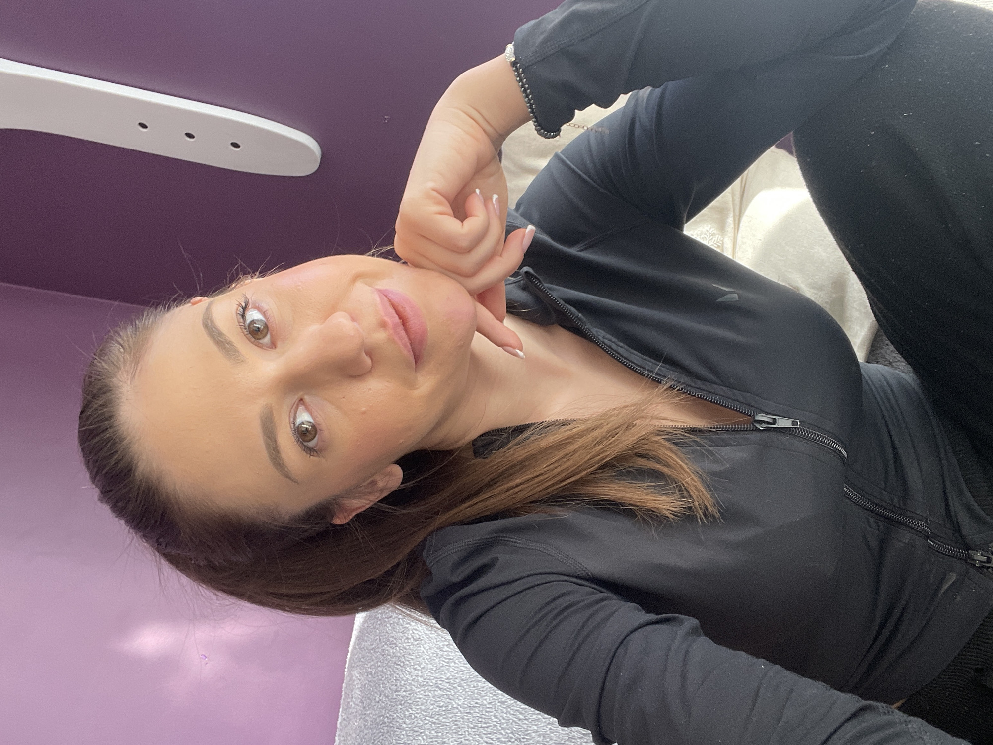 A woman with brown hair is positioned in the center of the frame, wearing a black zip-up jacket and black pants. Her head is slightly tilted, and her hand is near her face. The background is a blend of purple and gray, possibly walls or furniture. In the upper left corner, a white, elongated object with holes suggests a lighting fixture. The location is indoors, potentially in Sofia, Bulgaria, but no specific landmarks or indicators within the image confirm this.