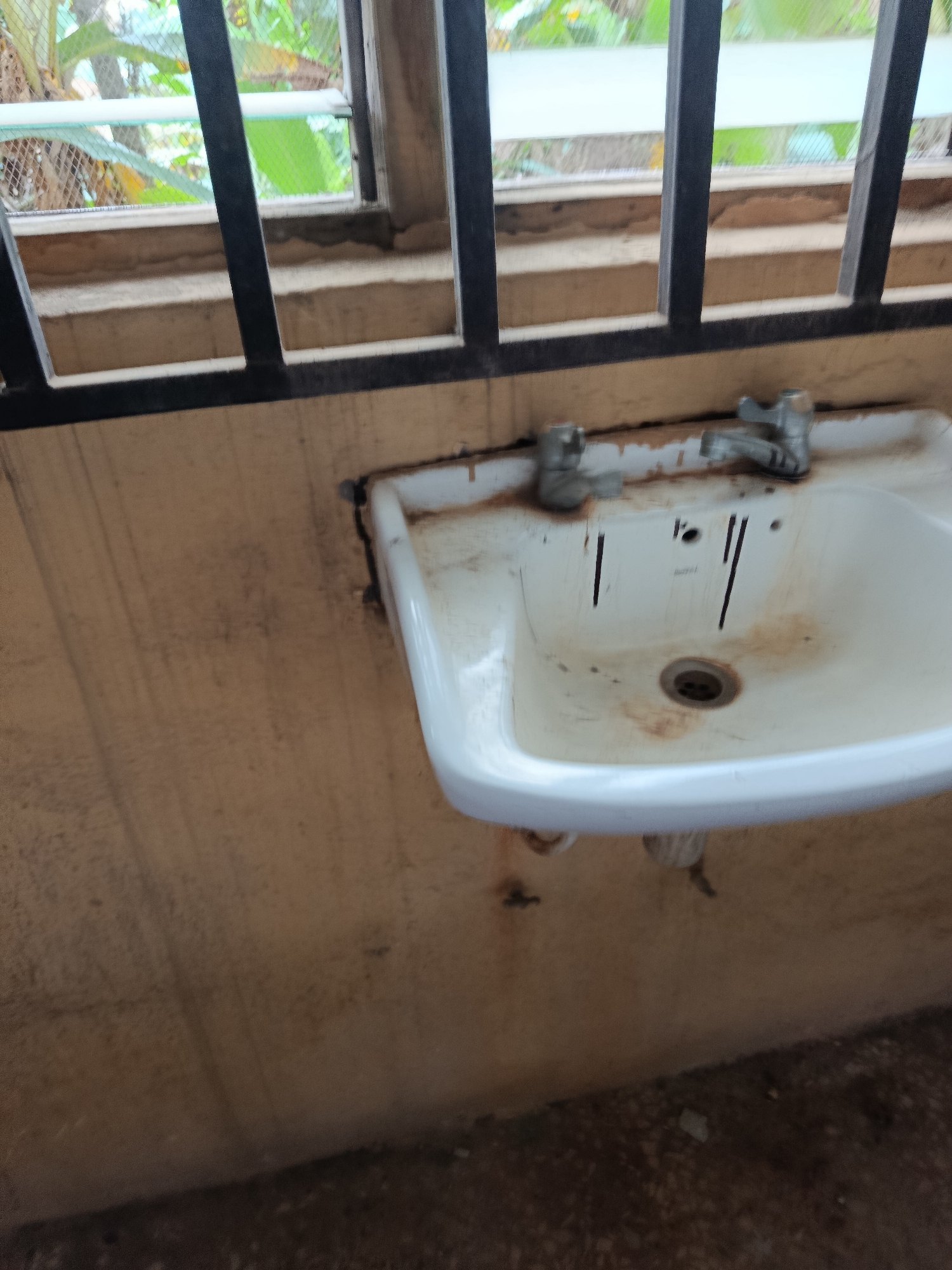 This is a medium-shot, eye-level, vertical photograph of a dirty white sink with two silver faucets. The sink is mounted on a light brown wall, which has streaks and stains running down it. Behind the sink, a window is visible with black metal bars in front of it. Through the window, greenery and trees can be seen, suggesting an outdoor or semi-outdoor setting. The floor in the foreground is a dark, uneven surface, possibly dirt or concrete, with some scattered debris. The overall impression is one of disrepair and neglect. No people are visible in the image. There is no discernible text. The lighting suggests it might be daytime, but the weather is not clear.