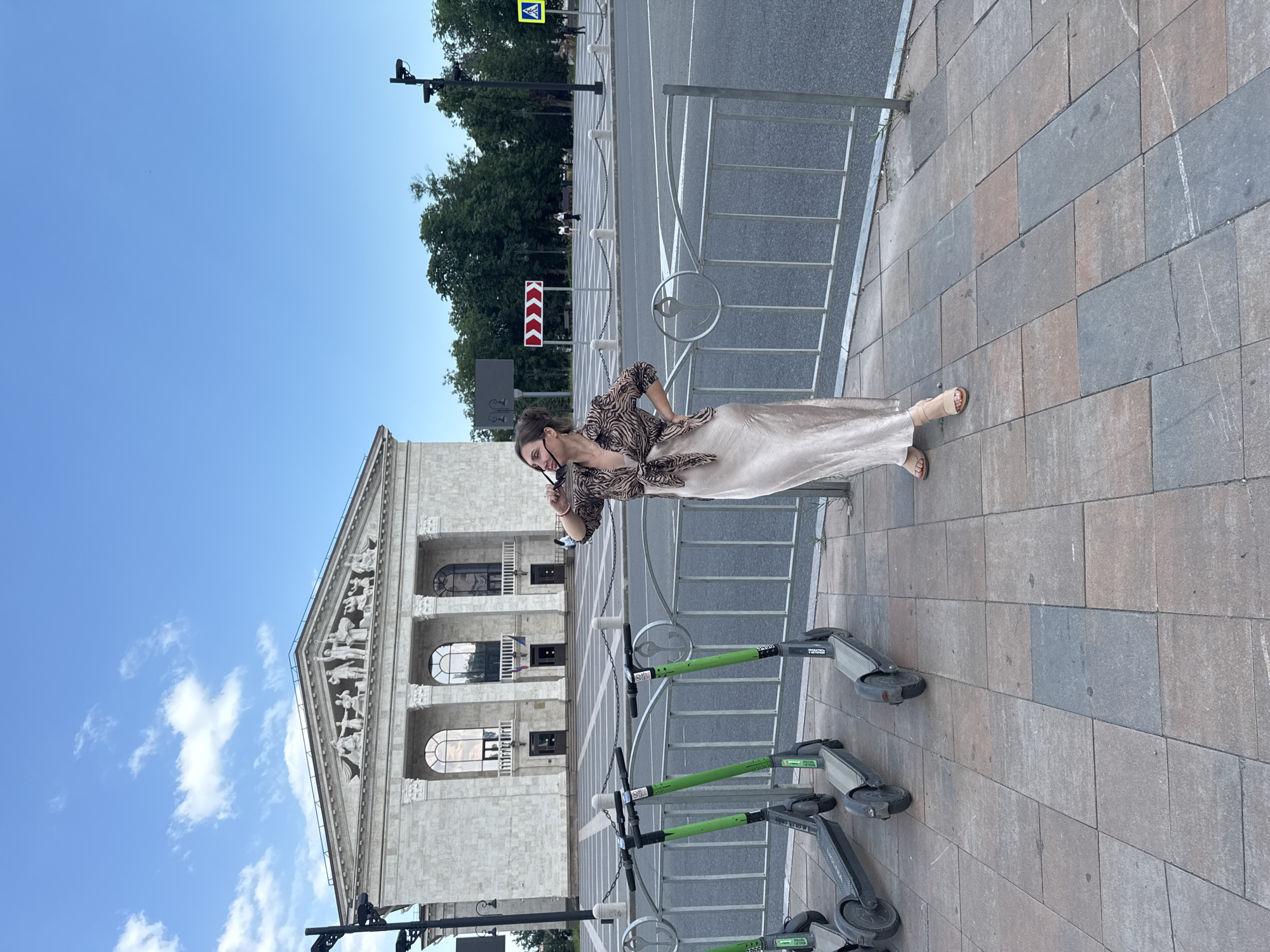 A young woman stands on a sidewalk in front of a large, ornate building. She is wearing a patterned blouse and a long, flowing skirt. She is holding her sunglasses and looking down. The building appears to be a government or cultural institution, with classical architecture featuring columns and arched windows. The sidewalk is paved with rectangular stones. To the left of the woman, there are several electric scooters parked. In the background, there is a street with traffic and trees. The sky is blue with a few white clouds, suggesting it is daytime and the weather is fair. The scene is set in Mariupol, Ukraine. The woman's posture and facial expression suggest she is thoughtful or perhaps surprised. Visible text on the scooters includes the brand name "Lime" and some numbers. A road sign with red and white arrows indicating a detour is also visible.
