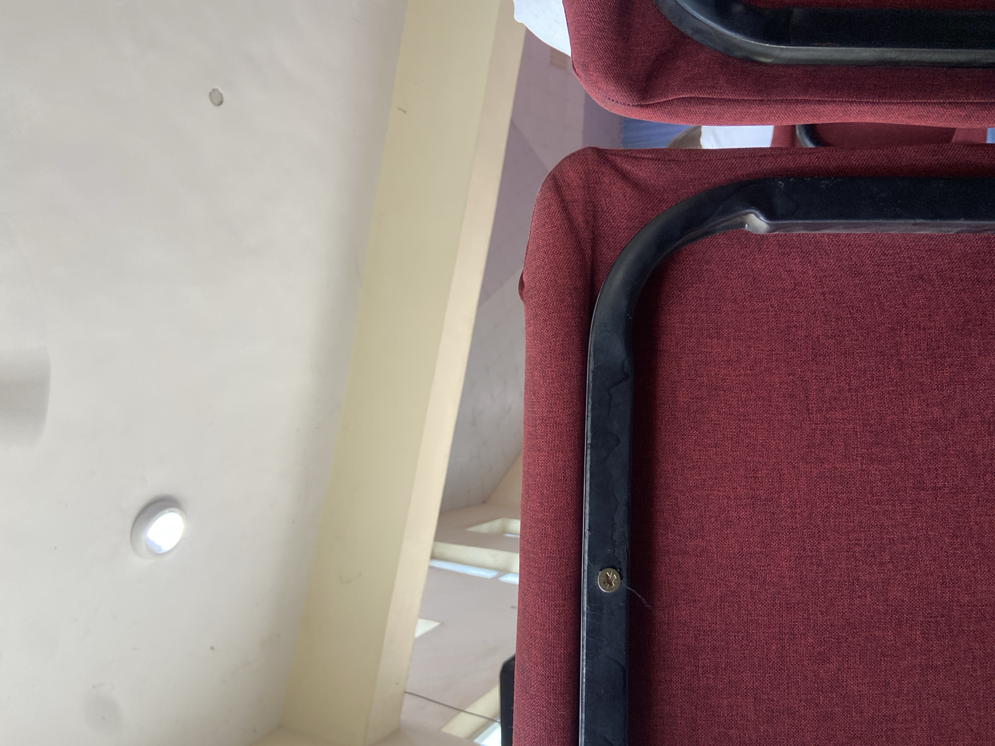 This image presents an indoor scene, observed from a low angle, within a structure in Mararaba, Nigeria. The foreground is occupied by the upper backs of two maroon-red upholstered chairs with black metal frames, seen from behind. A prominent gold-colored screw with a Phillips head is visible on the top horizontal bar of the left chair's frame. Above the chairs, a light yellow architectural beam or wall segment runs horizontally across the midground. The background reveals a white ceiling featuring a brightly illuminated circular recessed light fixture in the upper left, and a second, unlit, similar fixture positioned further right. Beyond the yellow beam, geometric architectural elements suggest windows or openings, allowing daylight to enter, creating patterns of light and shadow against a distant wall or sky. No individuals, specific actions, or interactions are visible.