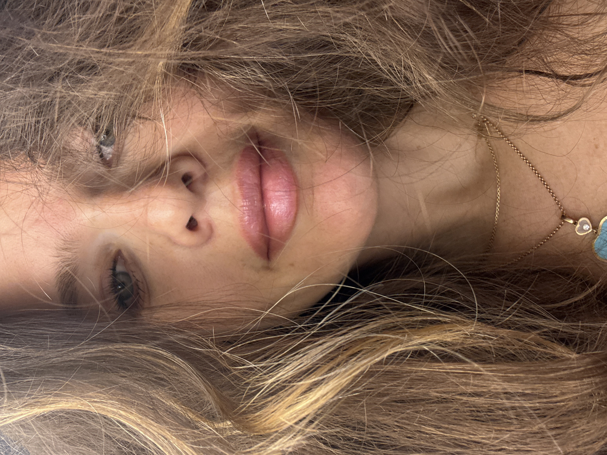Close-up, horizontally oriented shot of a person's face surrounded by hair. The face is slightly rotated, with the nose and mouth centrally located. One eye is clearly visible with dark lashes, and the other is partially obscured by hair. The skin tone is light. The lips are full and pink. A delicate gold necklace with a pendant is visible on the neck. The hair is a mix of light brown and blonde tones. The location is Unknown, Unknown.