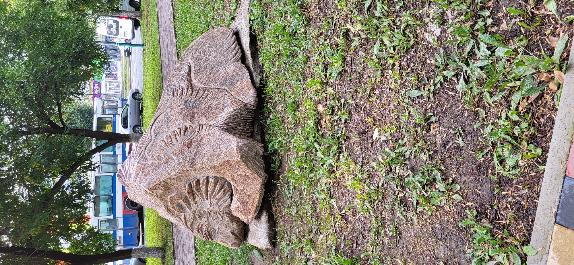 The image shows a carved stone sculpture resembling a bear's head, situated on a grassy area next to a sidewalk in Oskemen, Kazakhstan. Behind the sculpture, a blue and white bus is visible on a street, along with other vehicles and buildings. The sculpture itself is made of a light brown, speckled stone and is intricately carved with details suggesting fur and facial features. It rests on a flatter stone base. The surrounding environment includes trees with green foliage, grass, and a paved pathway.