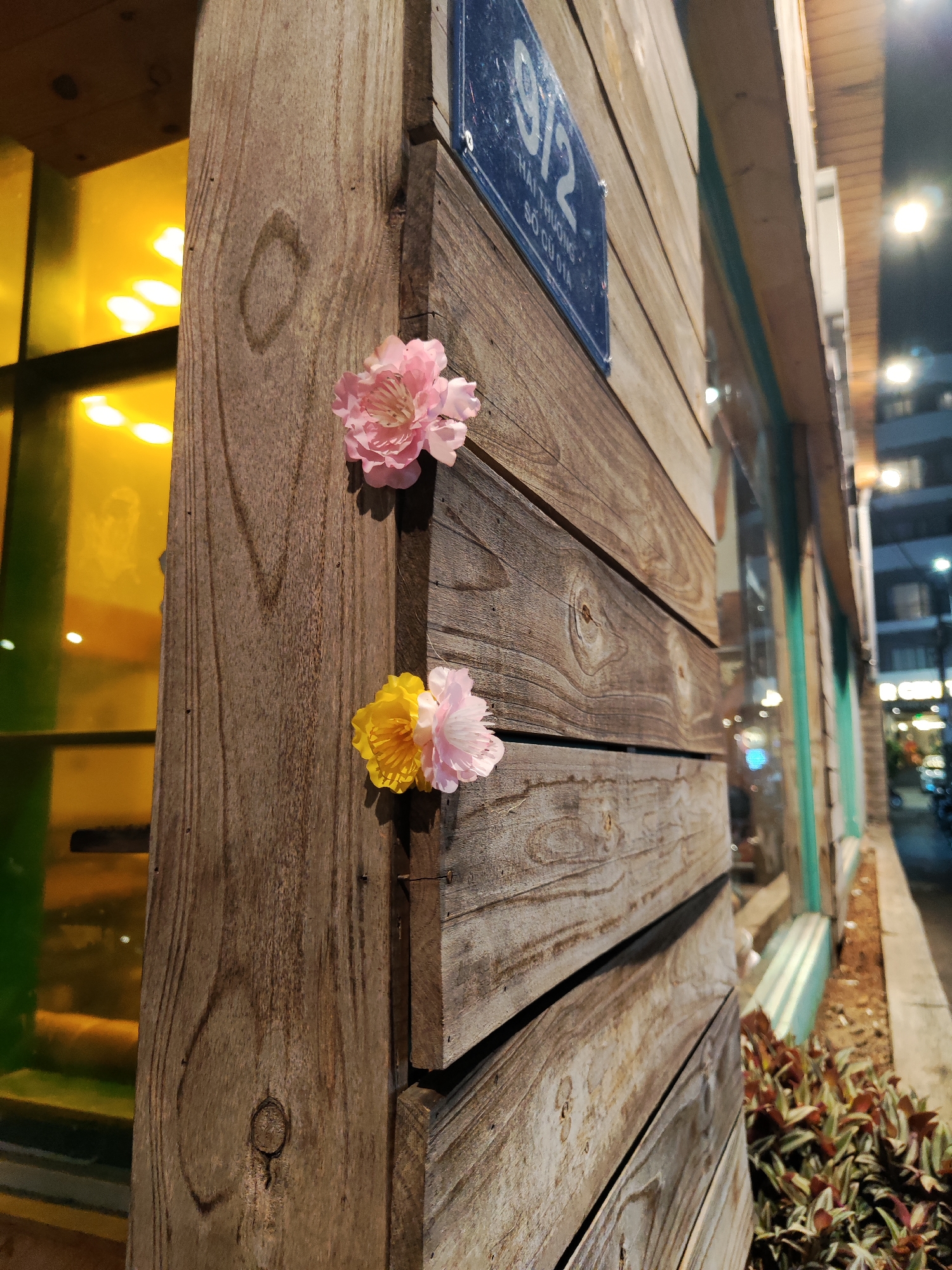 The image shows the exterior of a building at 9/2 Hai Thuong, So Cu Dia, Cam Ly - Đà Lạt, Vietnam. The building facade is constructed of weathered wooden planks. Two artificial flowers, one pink and one yellow-and-pink, are affixed to the wood. A window with yellow and green panes is visible to the left. In the background, a street scene is visible at night, with buildings and lights suggesting an urban environment. Plants line the base of the building's exterior wall.