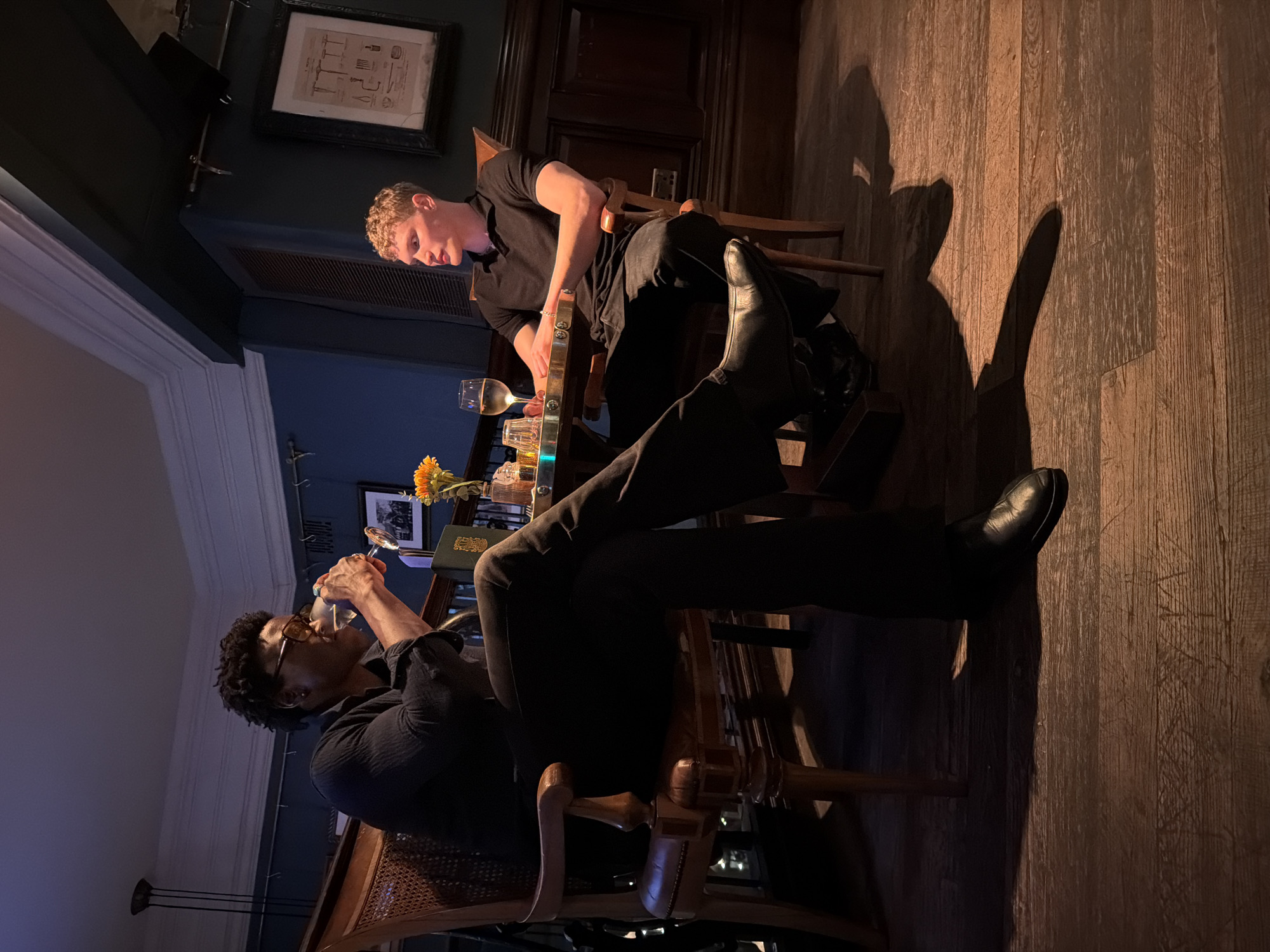 The image, rotated 90 degrees counter-clockwise, presents a dimly lit indoor scene featuring two individuals seated in what appears to be a cozy bar or restaurant, likely in London.
In the foreground, on the right side of the frame, a person with dark, curly hair and glasses, wearing a dark long-sleeved shirt, is seated in a wooden chair with a cane back. They are holding a clear glass or bottle to their mouth, taking a drink, with their left leg crossed over their right. A black boot is visible on their right foot.
Further back and to the left, another person with short, light-colored hair, wearing a dark polo shirt and dark trousers, is also seated in a wooden chair. They are leaning slightly forward, with one arm resting on a table or bar surface. A shiny bracelet adorns their left wrist.
Between the two individuals on the table, a wine glass containing white wine is visible, alongside a small vase with a yellow flower, a smaller framed picture, and a green-covered book with the word "GREAT" faintly visible on its spine. Above the light-haired person, a larger framed picture, possibly an old illustration, hangs on the dark blue wall. The ceiling features white molding. The floor is made of light-colored wooden planks, displaying strong shadows cast by the figures and furniture due to the warm, dim artificial lighting. The atmosphere appears relaxed and intimate, suggesting an evening social gathering.