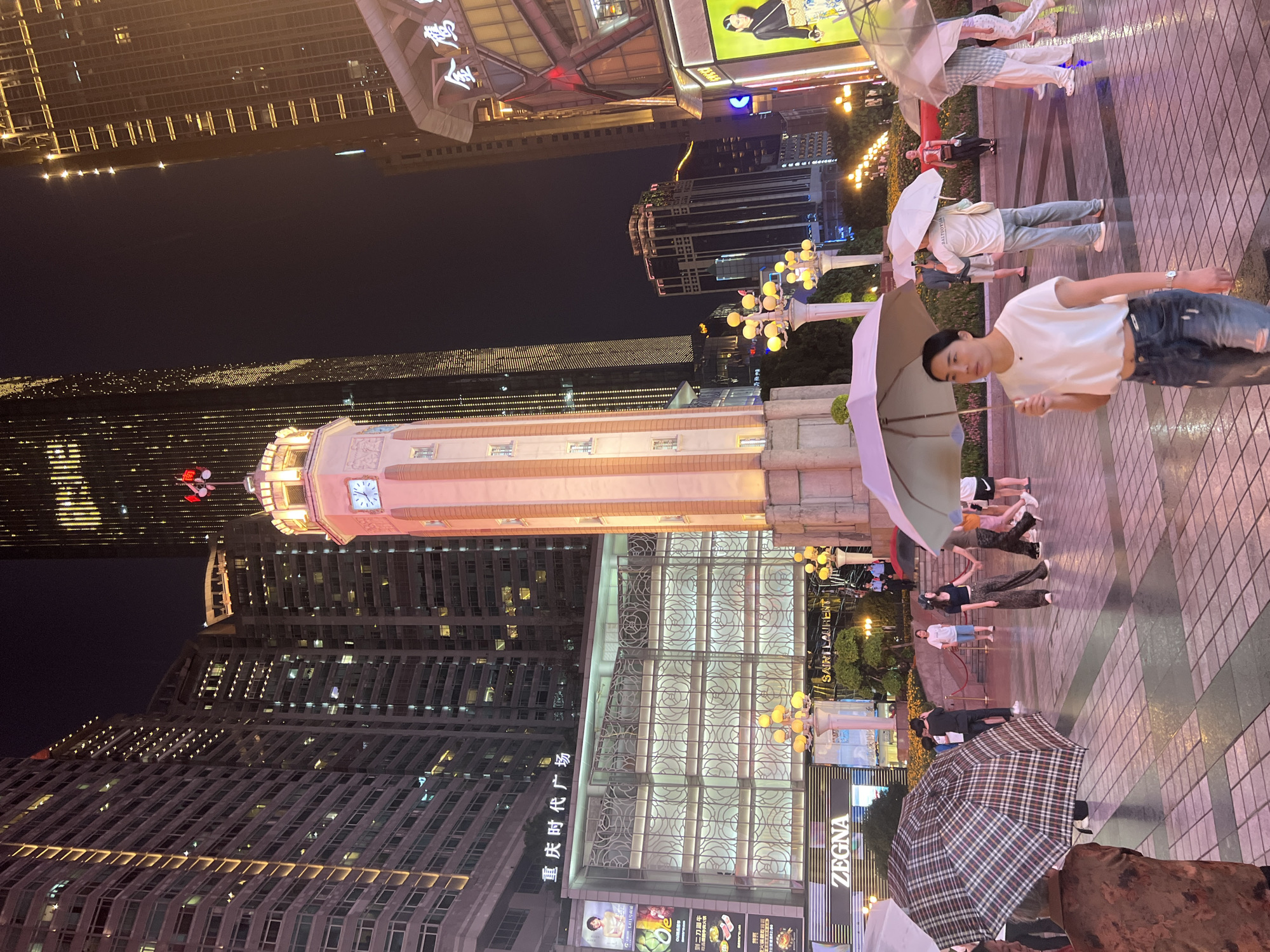 The image captures a nighttime street scene in Yuzhong District, China, characterized by towering illuminated buildings and a prominent clock tower. Several people are present, many holding umbrellas, suggesting it might be raining or has recently rained as the ground appears wet and reflective. In the foreground, a woman in a white crop top and ripped jeans walks forward, holding a light-colored umbrella. Behind her, other individuals stroll, some in groups, under umbrellas of various designs. A large checkered umbrella is held by someone on the left, and a light pink umbrella is visible further back.
The environment is urban, with a grand clock tower as a central focal point, adorned with lights and a red flag at its peak. Adjacent to the clock tower is a modern building with a glass facade displaying a floral pattern and featuring signs such as "ZEGNA" and "SAINT LAURENT". The surrounding skyscrapers are brightly lit, with many windows illuminated, creating a vibrant cityscape. Street lamps with multiple glowing orbs line the walkway. In the background, more buildings of varying heights and architectural styles contribute to the dense urban landscape. The overall impression is of a bustling commercial and pedestrian area at night.