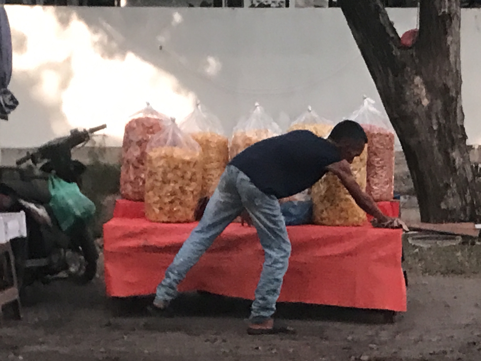 In Lhokseumawe, Indonesia, a man wearing a dark shirt and light-colored jeans leans over a red table displaying five large bags of potato chip snacks. The vendor is near a tree and a parked motorbike, with a white wall visible in the background. The ground is dark and appears to be an unpaved area.