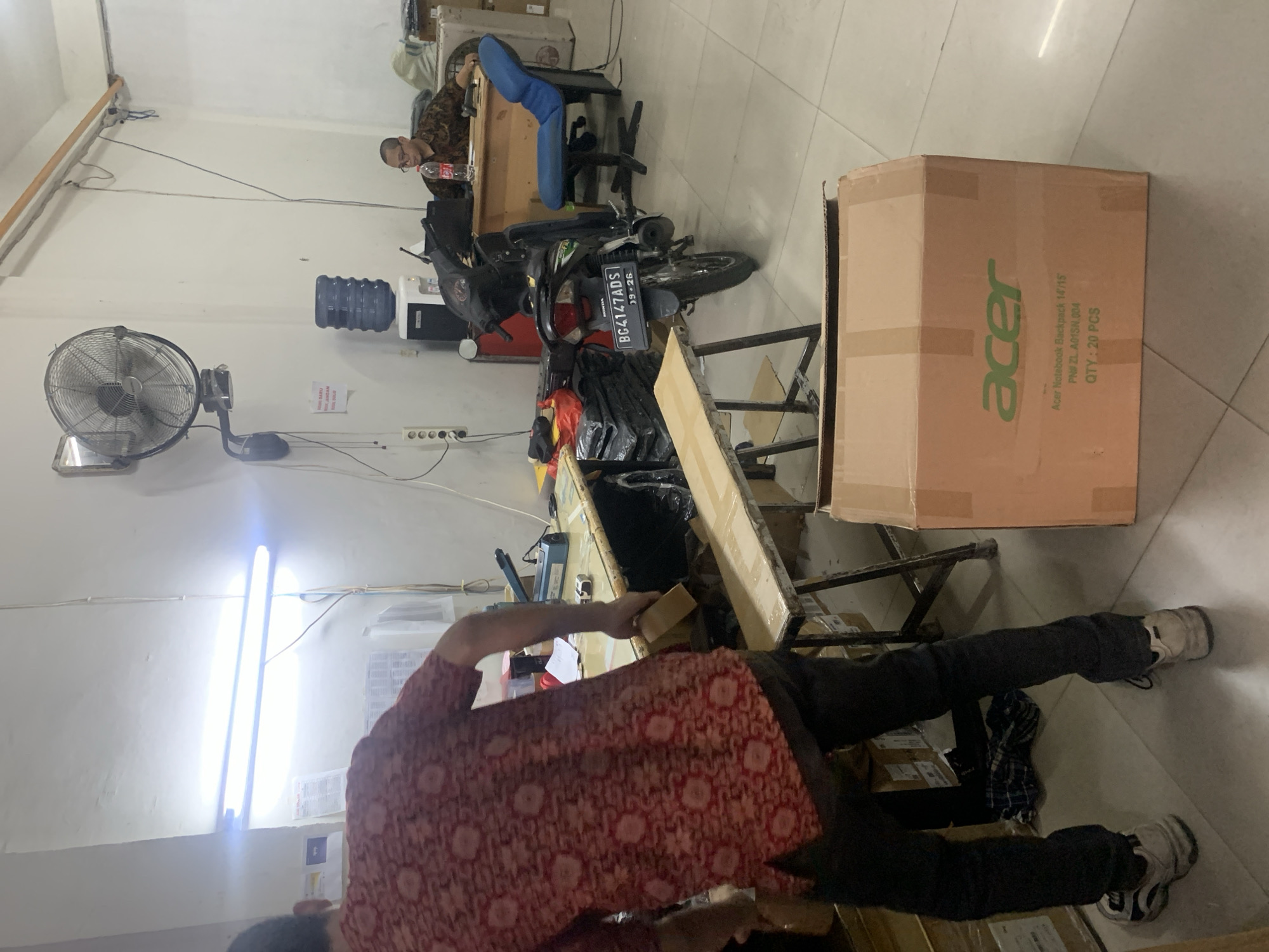 The image depicts an indoor scene, likely a warehouse or storage area, located in Palembang, Indonesia. In the foreground, a man wearing a red and white patterned shirt and dark pants is standing and holding a light brown box. His back is partially to the camera. In the background, another man is seated at a desk, wearing glasses and a dark patterned shirt. He appears to be working on a computer. A blue office chair is situated behind him. A motorcycle is parked near the desk. There are several boxes stacked around the room, including a large brown box with "acer" logo prominently displayed. A water dispenser with a large blue jug is visible on a stand. The room is illuminated by bright fluorescent lights. The walls are white and appear to be concrete. The floor is tiled. The overall impression is a functional, utilitarian space.