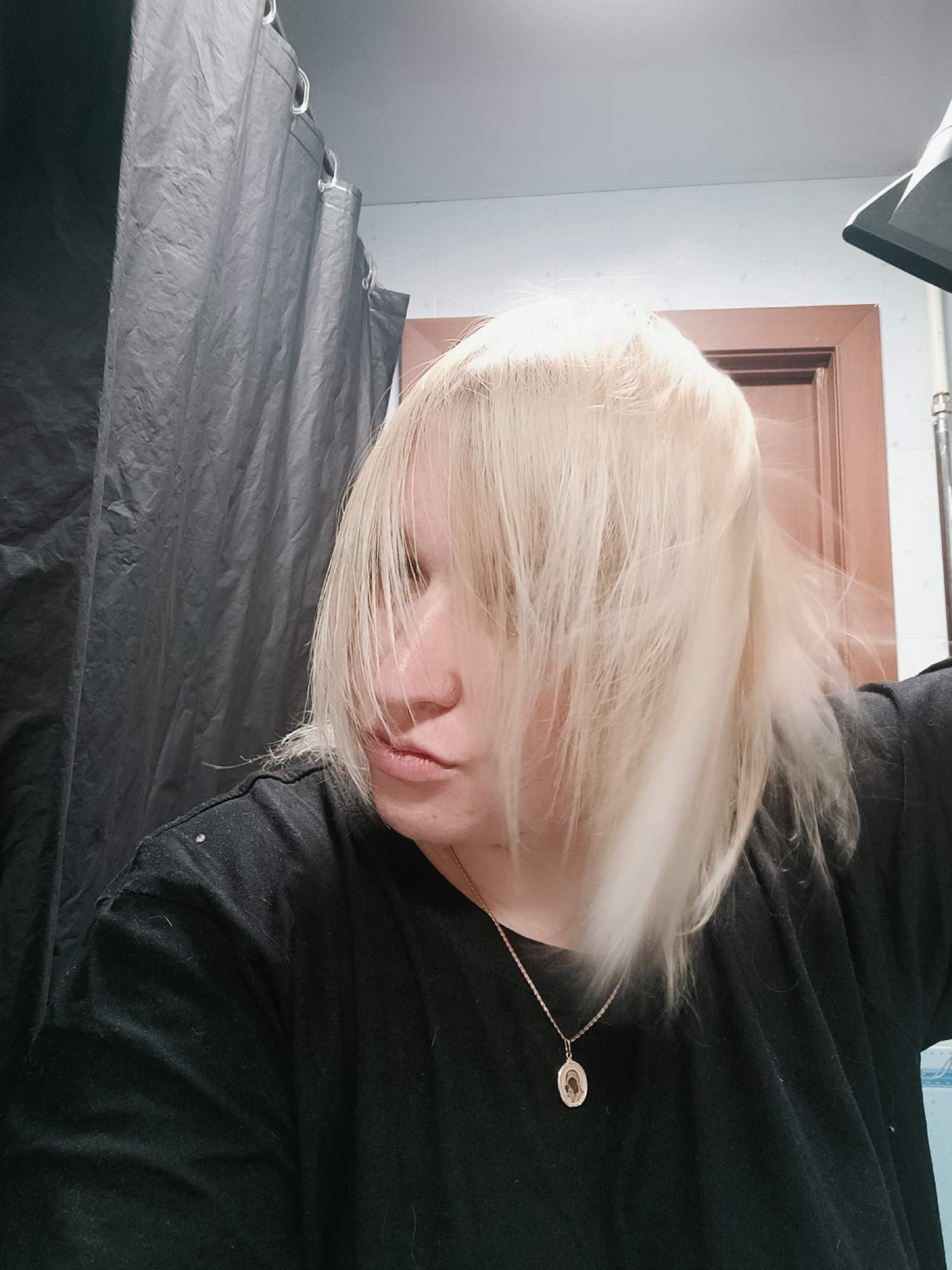 A medium shot depicts a person with platinum blonde, shoulder-length hair, wearing a black t-shirt and a gold necklace with a pendant. The person is looking slightly to the left with a neutral expression. The hair is covering part of their face. Behind the person is a black shower curtain on the left side of the frame. To the right of the person is a light-colored wall, a brown door, and a bathroom fixture. The location is assumed to be inside a bathroom in Chaikovsky, Russia.