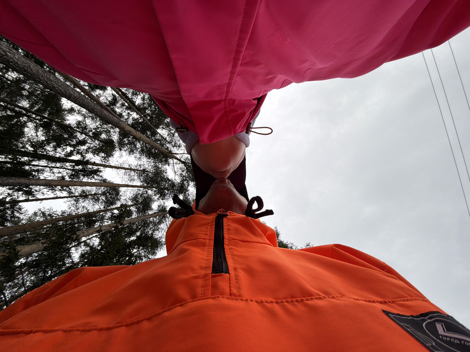 Two people are positioned close together, likely kissing or about to kiss. The person at the top is wearing a pink jacket, while the person below is wearing an orange jacket. The background includes trees, likely in a wooded area. The location may be the city of Chaikovsky, Russia, as indicated by text on a patch on the orange jacket. The sky is overcast.
