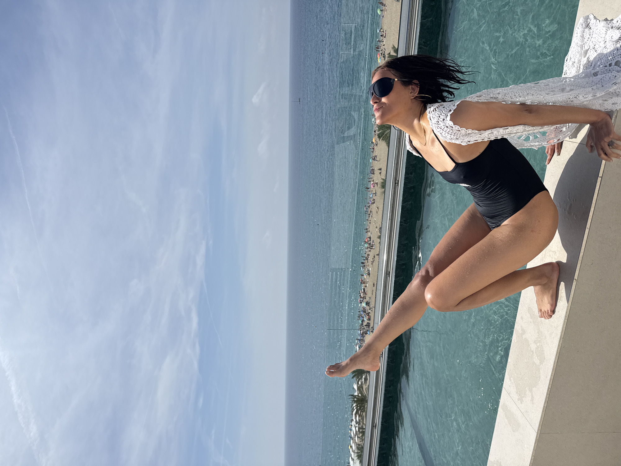 An adult female is positioned on the edge of an infinity pool, with a panoramic view of the Mediterranean Sea and a beach in Calella, Spain. She wears a black one-piece swimsuit, black sunglasses, gold hoop earrings, and a thin gold necklace. A white, open-weave crocheted cover-up drapes over her left shoulder and down her back. Her dark hair appears wet, and water droplets are visible on her skin, particularly her legs.
She is leaning back, supporting herself with both hands on a light beige concrete ledge adjacent to the pool. Her head is tilted upwards towards the sky, and her eyes are closed or looking up under the sunglasses. Her right leg is bent with her foot flat on the ledge, while her left leg is extended upwards and slightly forward, bent at the knee.
The pool water is clear, turquoise, and gently rippled. Beyond the transparent glass barrier of the infinity pool, a wide sandy beach is visible, densely populated with numerous people, many sitting under colorful beach umbrellas. Several white sunshades or small structures are present at the back of the beach, with palm trees interspersed. The calm, deep blue sea extends to the horizon under a bright, light blue sky featuring scattered wispy clouds and faint contrails. A single small sailboat is discernible in the distant sea.