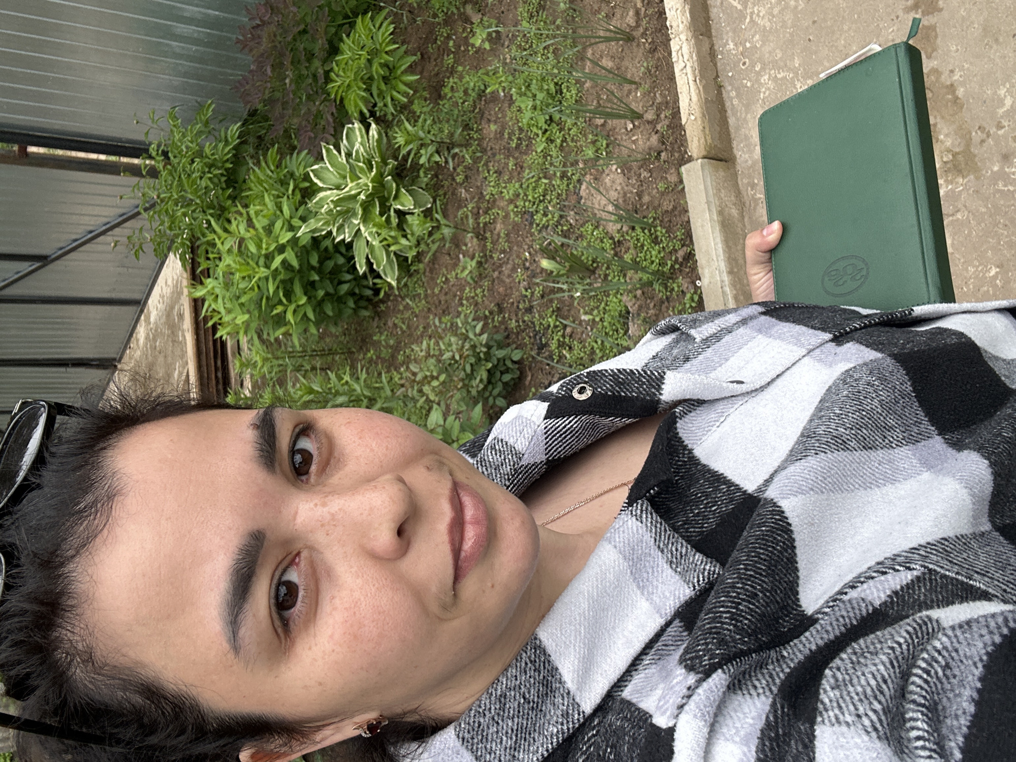 A woman is depicted in a self-portrait or close-up shot outdoors, looking directly at the camera with a neutral expression and a slight smile. She has dark hair, dark eyebrows, brown eyes, and visible freckles on her face. She is wearing a black and white plaid shirt, a thin gold chain necklace, and a small reddish earring in her left ear. Sunglasses are perched on top of her head.
In her right hand, she holds a dark green book or planner with a debossed logo on its cover, which appears to be a stylized "BB" or "22". The background consists of a garden or backyard setting, featuring various green plants, including a hosta with variegated leaves, and ground cover growing in soil. To the left, a metal fence structure is partially visible, and concrete or stone paving runs along the right side of the frame and further into the background. The scene is illuminated by soft, natural light, suggesting it is daytime, possibly overcast.