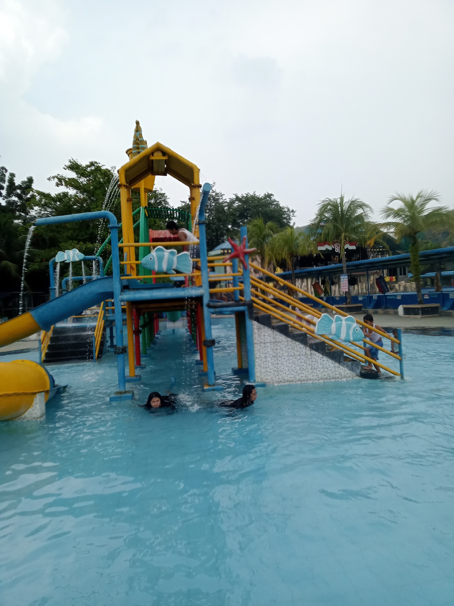 An outdoor water park with a brightly colored slide structure is depicted. The slide is primarily yellow and blue with red accents and has multiple levels and stairs. Two children are visible in the clear blue water. One child is swimming in the foreground, while another is near the base of the slide structure. The water park appears to be located in Baru, Indonesia, as indicated by the presence of palm trees and tropical vegetation. The sky is overcast, suggesting a cloudy day.