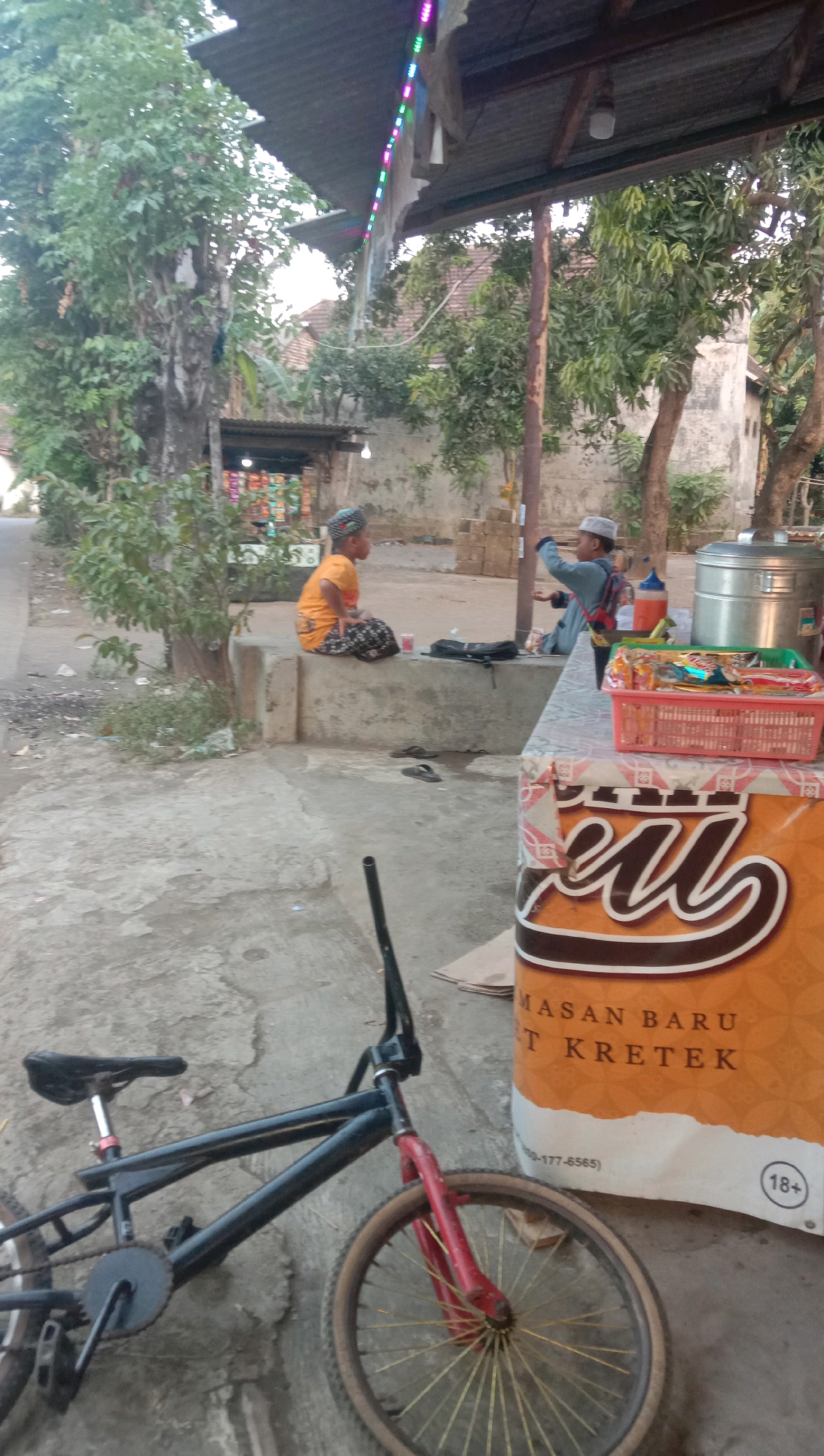 In the village of Ngempit, Indonesia, a BMX bicycle is parked on a concrete surface near a stall. The bicycle is black with red forks and golden spokes. To the right of the bicycle, a large orange banner with white text and a red and white patterned border is visible. The banner appears to advertise a product, with text that reads "MASAN BARU T KRETEK" and a phone number. An "18+" symbol is also present on the banner. Behind the stall, a woman in an orange shirt and patterned skirt sits on a concrete ledge, facing left. Further back, a young boy in a white cap and blue shirt is standing and appears to be taking a photo with his phone. A metal container and a red basket filled with snacks are on the stall. Trees and buildings are visible in the background, and colorful lights are strung across the top of the stall's roof.