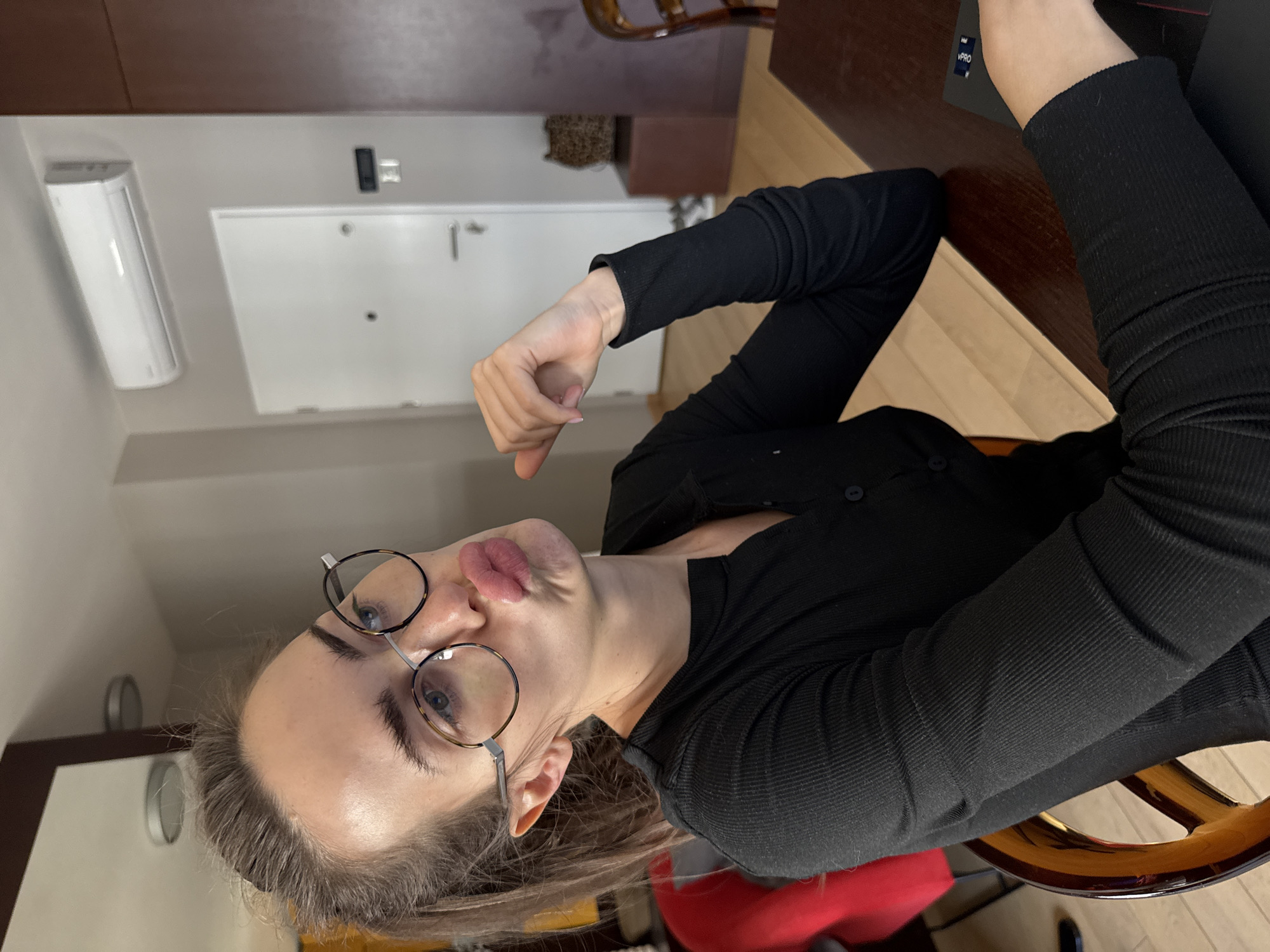 The image, rotated 90 degrees clockwise, captures a young adult female from the chest up, seated indoors at a dark wooden table. She looks towards the viewer with a playful expression, puckering her lips. She wears round, wire-frame glasses with tortoise-shell accents and a black, long-sleeved, ribbed top or cardigan. Her light-colored hair is visible around her face. Her left hand is raised slightly, with fingers loosely curled.
On the table to her right, a dark laptop is partially visible, bearing the text "Intel" and "vPRO". The background features light-colored walls and a light wooden floor. A white door with a visible handle is in the distance. To the left of the door, a white, wall-mounted air conditioning unit is installed. Above the door, a small dark rectangular object and a lighter switch-like panel are visible on the wall. Part of a red chair cushion and a wooden chair frame are also discernible. The scene suggests a casual daytime setting within a modern apartment or office in Warsaw, Poland.
