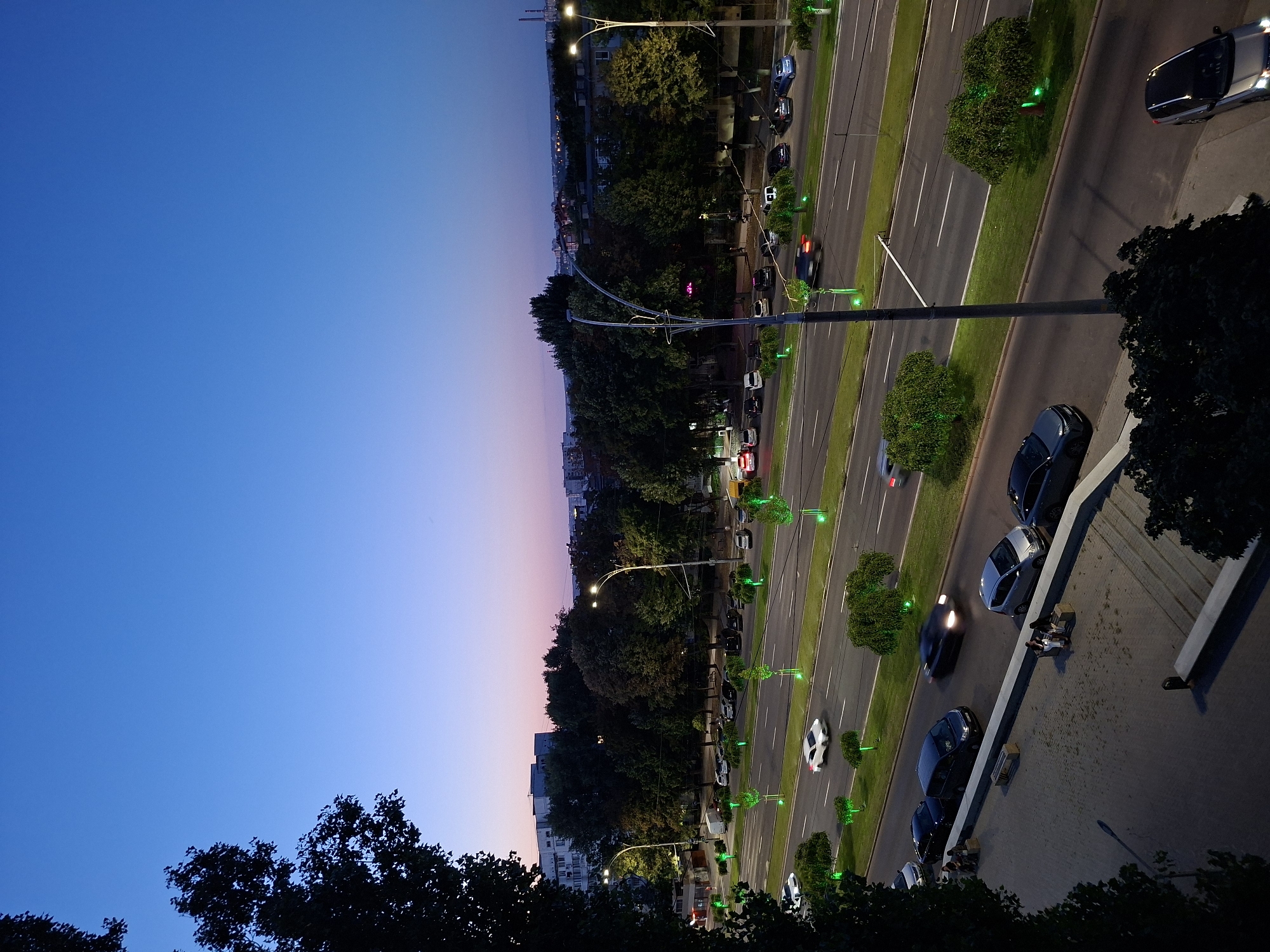 The image depicts an elevated view of a wide, multi-lane road in Chișinău, Moldova, during twilight. The sky transitions from a deep blue at the top to lighter shades of purple and pink near the horizon. Along the roadside, numerous trees are illuminated with green lights, creating a vibrant contrast with the darkening sky and road. Streetlights with curved arms are visible, also illuminated.
Several cars are in motion on the road, including a white sedan in the middle lane, a black sedan in the right lane, and a silver hatchback in the left lane. Parked cars are visible on the side of the road further up, and some cars are driving in the opposite direction. On the left side of the road, there is a pedestrian area with a few individuals sitting on benches. A concrete barrier separates the pedestrian area from the road. In the background, buildings and trees form the urban landscape. The overall scene suggests a well-lit urban environment at the end of the day.