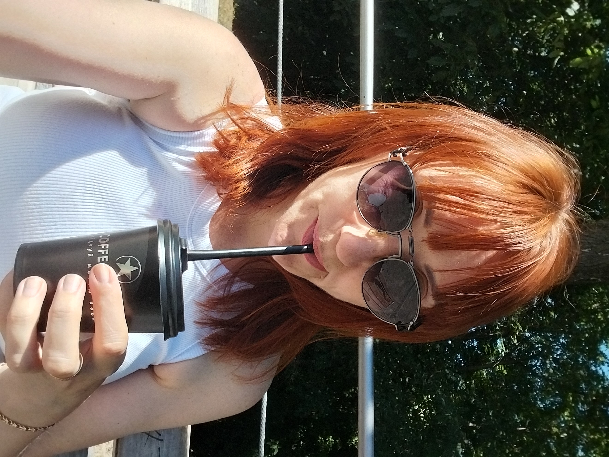 A woman with auburn hair and sunglasses is holding a black coffee cup and drinking from it with a straw. She is wearing a white ribbed tank top and is sitting on a bench outdoors. The background consists of green trees. The coffee cup has the word "COFFEE" and a star logo printed on it.