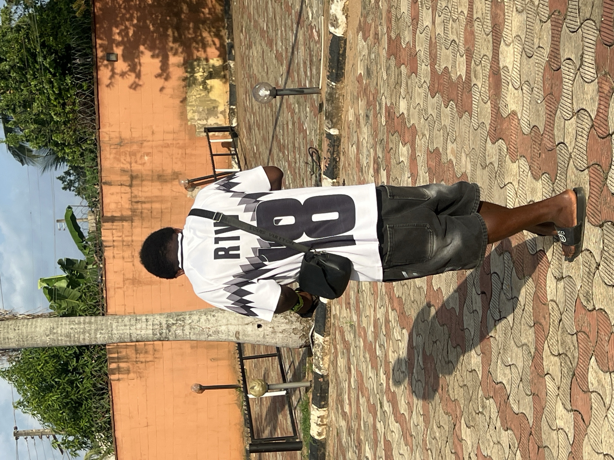 An outdoor, full-length shot shows a person from behind, standing or walking on a patterned tiled ground. The person, an adult male with dark skin, wears a white short-sleeved shirt featuring black and grey geometric designs and black denim shorts. A black cross-body bag is slung over their left shoulder, and they are wearing black slide sandals. On the back of the shirt, the word "RIVA" is visible above the large number "80". The strap of the cross-body bag faintly displays "LOUIS VUITTEN," and a "GUCCI"-like interlocking G logo is on the right slide sandal.
To the left of the person stands a tall, slender palm tree. The background features a light orange-brown wall topped with barbed wire and lush green foliage, suggesting a secure boundary. Above the foliage, patches of pale blue sky are visible. The ground is paved with interlocking tiles in wavy patterns of beige, brown, and terracotta. Two small lamp posts or decorative bollards are visible along the wall. The distinct, elongated shadow cast by the person and the palm tree indicates a sunny day, likely late morning or afternoon. The tropical vegetation and bright conditions align with the specified location context of Nkpor, Nigeria.