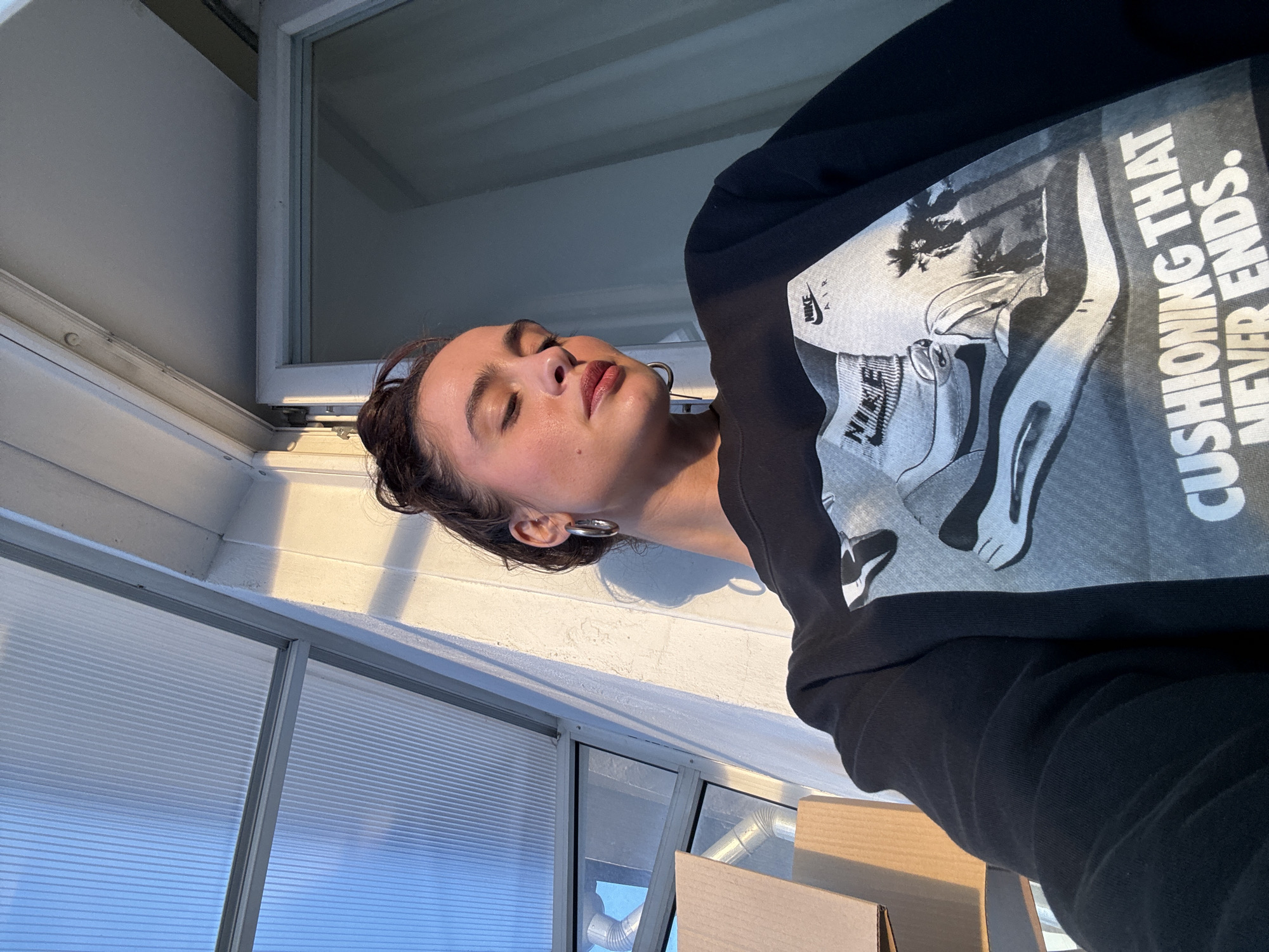 A medium shot captures a fair-skinned woman indoors, facing right, with her eyes closed. She is wearing a black sweatshirt featuring a black and white graphic of a Nike Air sneaker and the text "CUSHIONING THAT NEVER ENDS." She is wearing a hoop earring in her left ear.
The woman is positioned in front of a window with light streaming in. Behind her is corrugated plastic and a white painted wall. Parts of cardboard boxes are visible.
There is no indication of the location being Krasnoyarsk, Russia in the image.