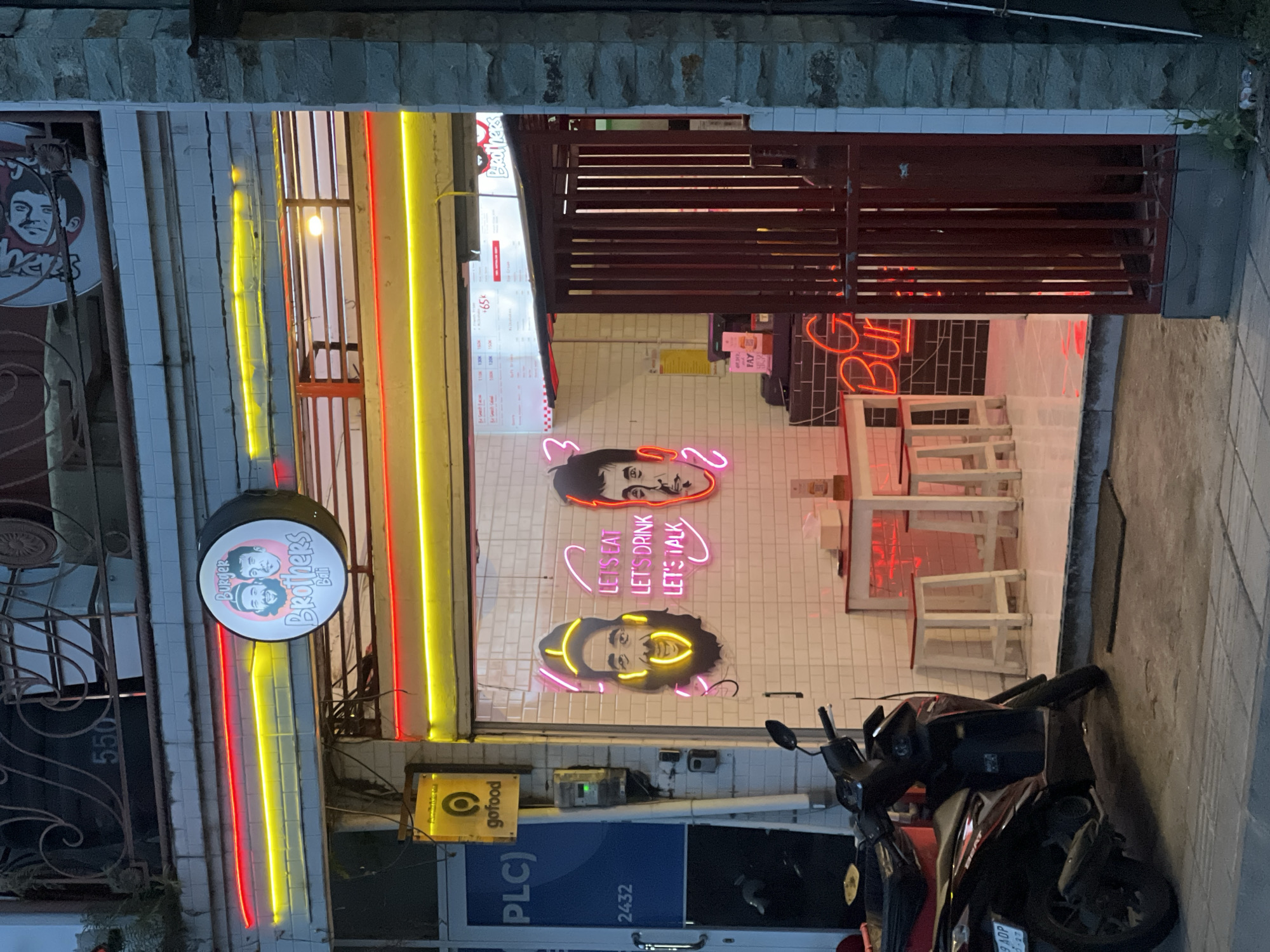 The image depicts the exterior of "Burger Brothers Bali," a restaurant located in Tibubeneng, Indonesia. The restaurant features neon signs of two men's faces and the text "LET'S EAT, LET'S DRINK, LET'S TALK" in a similar style. A circular sign on the left displays the restaurant's name and cartoon faces. A menu board is visible inside the restaurant. A GoFood sign is affixed to the building near the entrance. Two parked motorcycles are positioned in front of the establishment. The street number, 2432, and 550 are also visible.