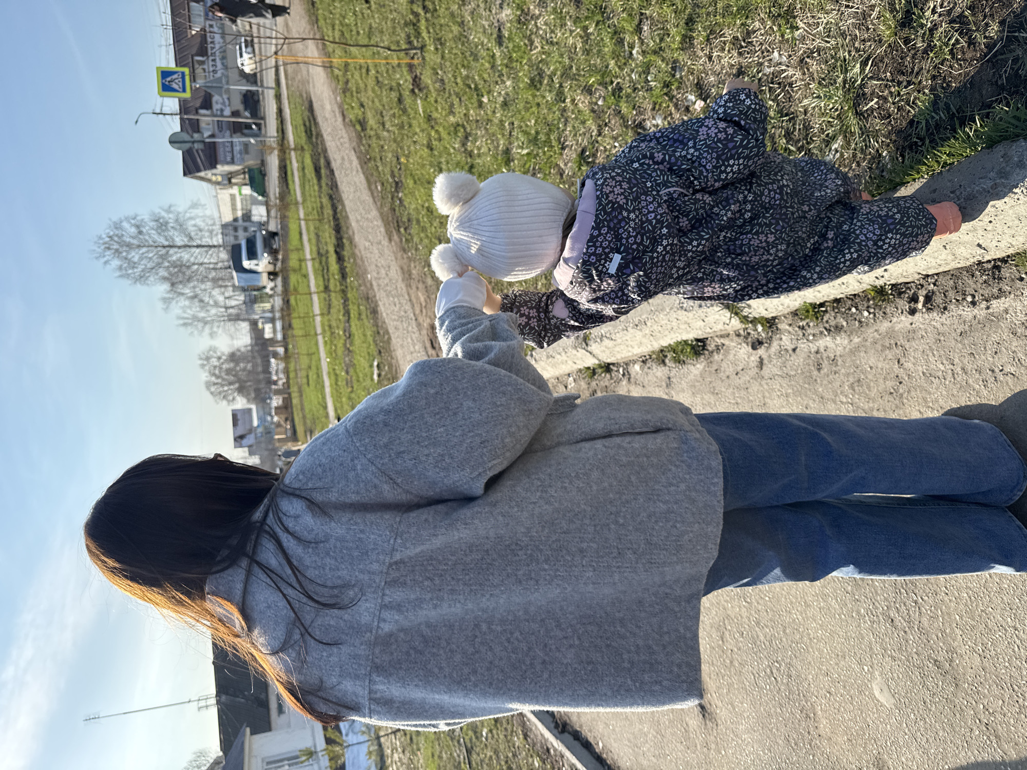A woman in blue jeans and a gray jacket stands on a sidewalk in the city of Chaikovsky, Russia, holding the hand of a young child wearing a white hat with pom-poms and a floral print jacket, who is reaching towards the curb. A street runs parallel to the sidewalk, with green grass and a ditch between the street and the sidewalk. A crosswalk sign and various buildings are visible in the background.