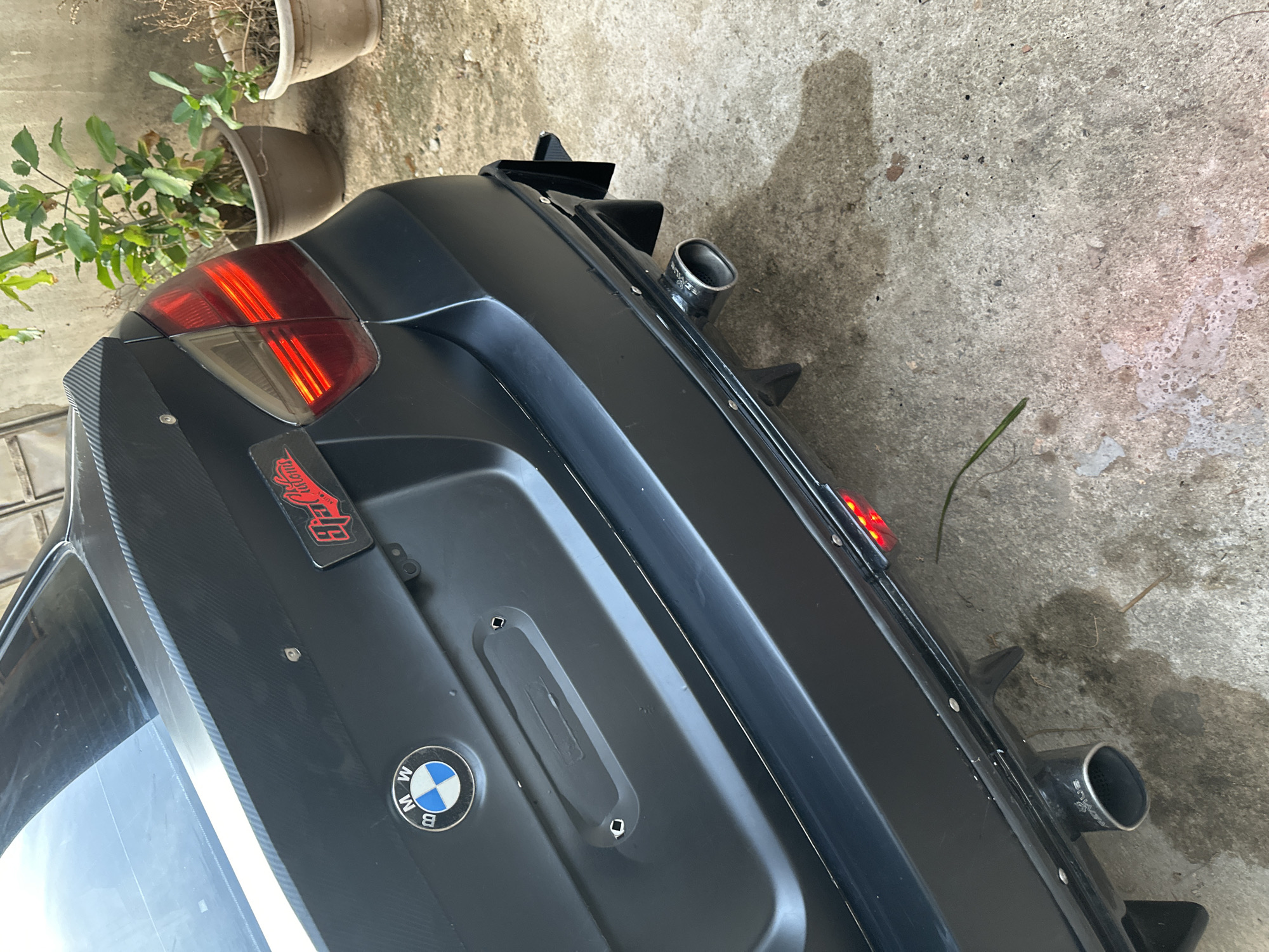 The image displays the rear-left quarter of a stationary, black matte BMW car, viewed from an elevated angle, on what appears to be an outdoor paved surface. The car features a heavily modified rear with a prominent black diffuser equipped with several sharp fins and dual exhaust pipes, one visible on each side, sporting a metallic finish. The left taillight of the car is visible, glowing red with distinct illuminated strips.
On the car's black trunk lid, a classic blue and white BMW emblem is clearly visible. Above it, a red sticker with the text "Hot Rod" and partially visible "Autom..." (likely "Automotive") is affixed. The trunk also has a trim piece with a carbon fiber pattern. The car is parked on a textured, light-colored concrete ground that shows some dirt and dried water stains. In the upper left background, several potted plants are visible against a light-colored wall, with one pot containing green foliage and another appearing dry. The scene is brightly lit, indicating daytime. No people or active events are present. The location context for this scene is Barden Yana, Nigeria.