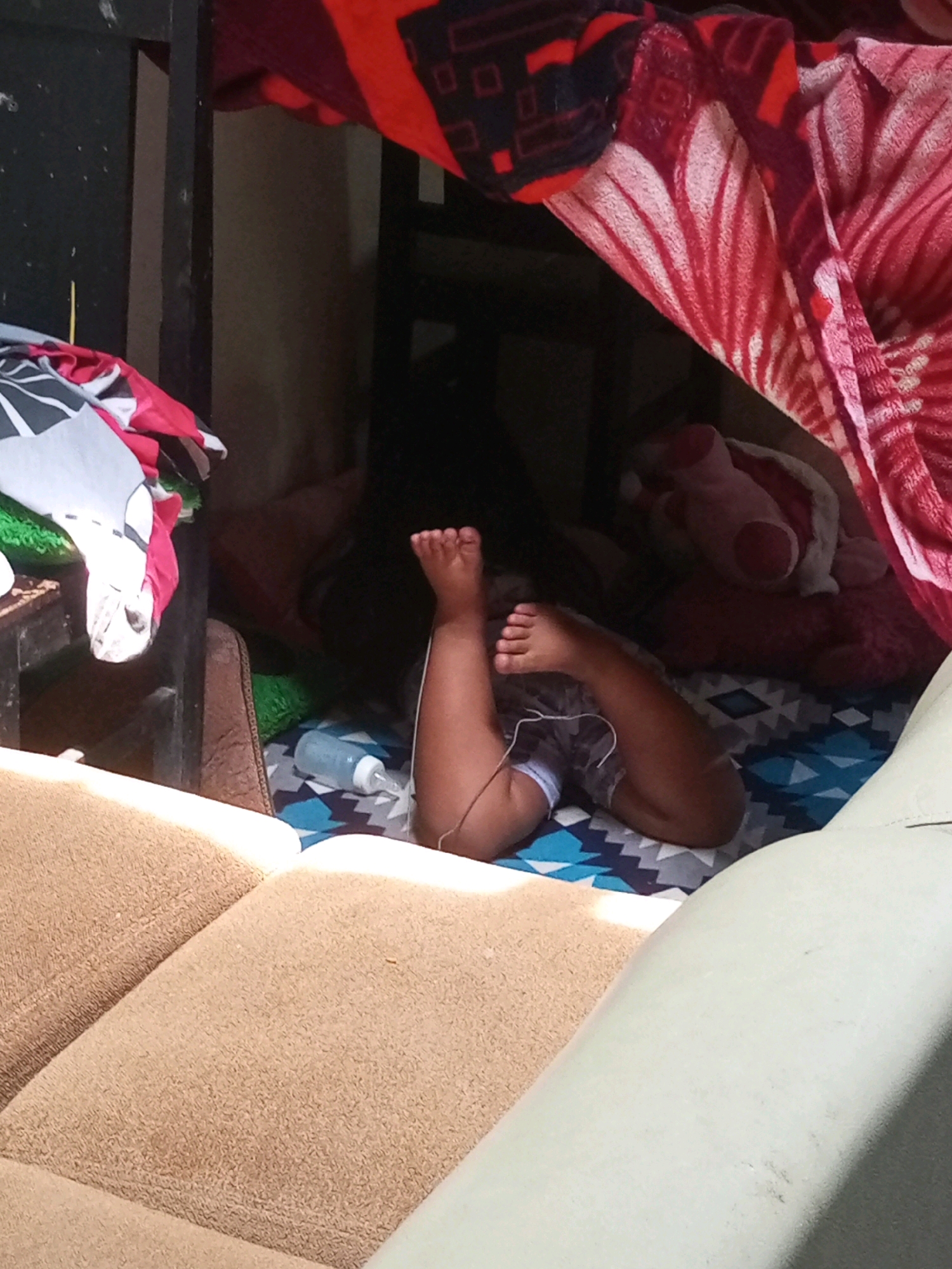 The image depicts a child's legs and feet protruding from under blankets and bedding. The child appears to be lying down, possibly sleeping or resting. A baby bottle with a blue cap is visible on a patterned mat near the child's legs. The scene is enclosed by various textiles, including a red and white patterned blanket and other colorful fabrics, suggesting a cozy or makeshift sleeping area. The background is dimly lit, hinting at an indoor setting or a shaded area outdoors. The overall impression is one of a relaxed and private moment, possibly in a home or a sheltered space. There is no visible text in the image.