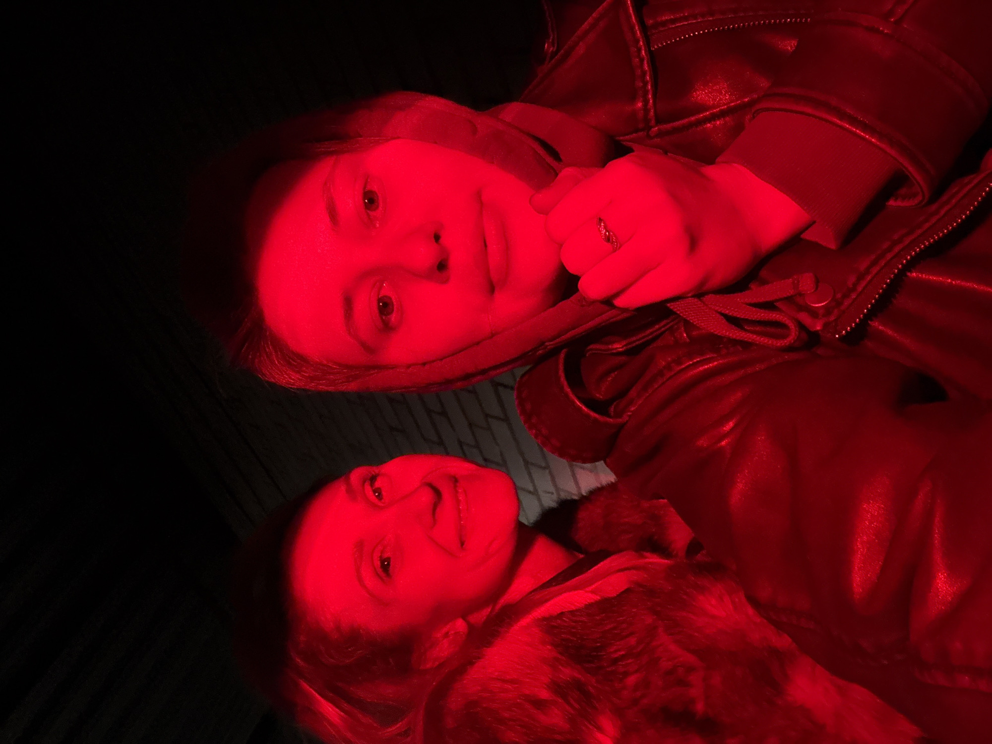Two individuals are captured in a dimly lit environment with a predominantly red lighting hue. The person at the top wears a leather jacket and a hoodie, with a hand visible near their face, adorned with a ring. The lower individual is wearing a fur collar jacket. The scene is possibly outdoors, with dark backgrounds and limited visibility, and could be illuminated by red lights such as tail lights. The location is the city of Chaikovsky, Russia.