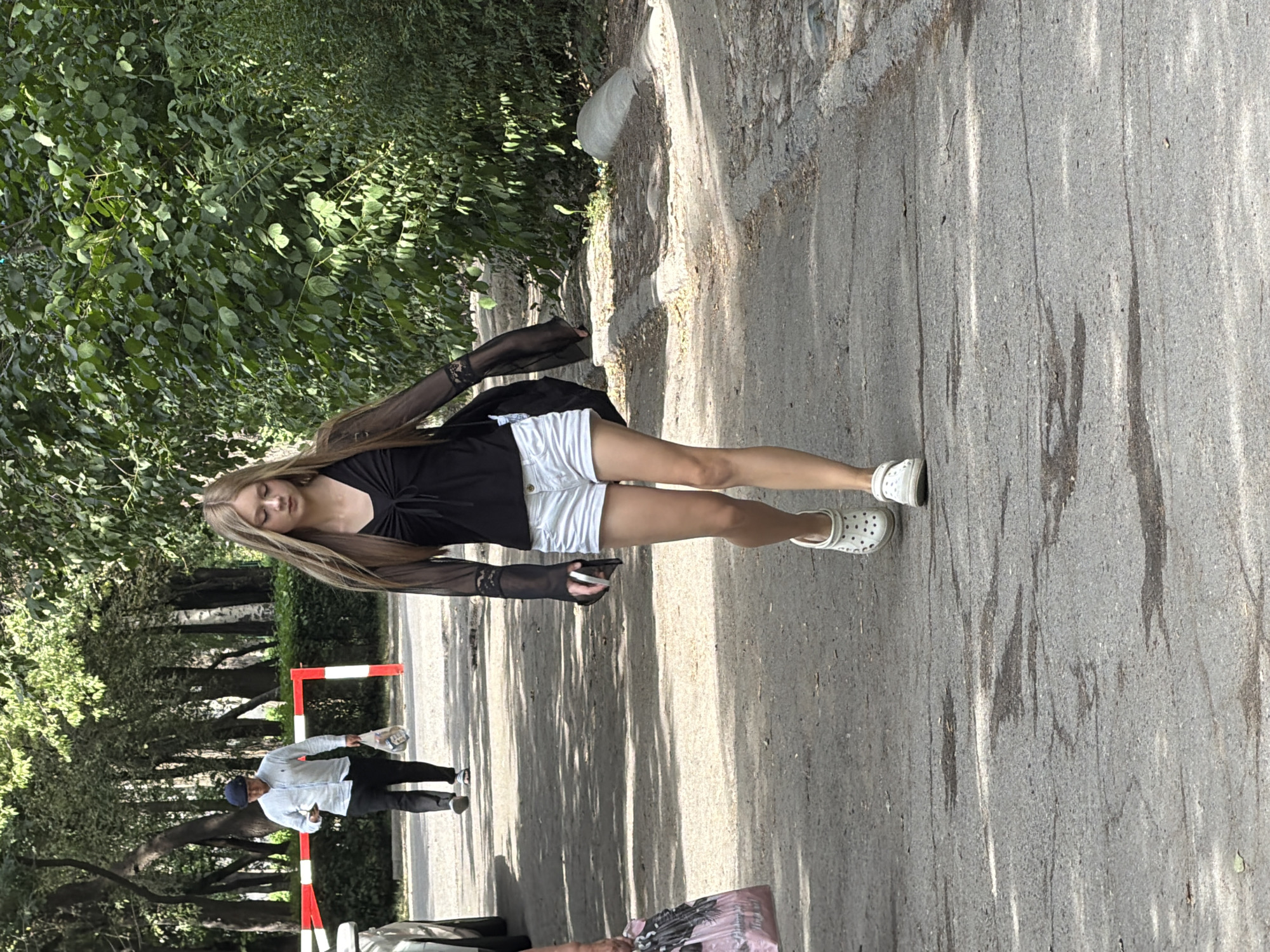 A young woman with long, blonde hair walks on a paved path in an outdoor setting. She is wearing a black, sheer long-sleeved shirt with bell sleeves and a V-neck, tied at the front, and white denim shorts. She has on white Crocs-style clogs. She is holding a mobile phone in her right hand. In the background, a man in a light blue collared shirt, dark pants, and a dark blue cap walks away from the camera, holding a plastic bag in his right hand and a phone in his left. He is walking past a red and white striped barrier. The environment is characterized by trees with green foliage, a paved pathway with some cracks and stains, and rough, rocky edges along the side. Sunlight filters through the trees, creating dappled shadows on the path.