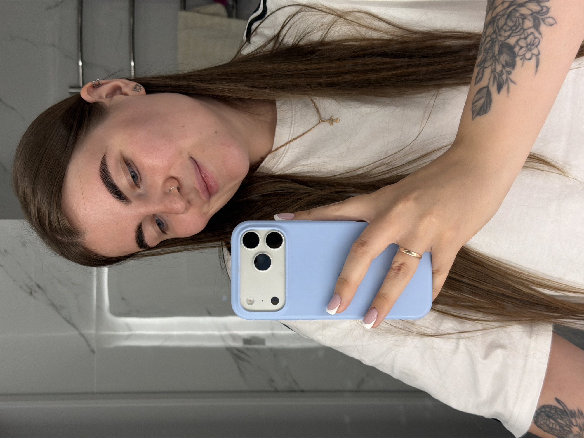 A woman is taking a selfie in a bathroom. She is holding a blue smartphone in her right hand. Her long brown hair is draped over her right shoulder. She is wearing a white t-shirt with a black stripe on the shoulder, and a gold cross necklace. She has a tattoo on her right forearm. The bathroom features a white marble wall. The location is likely Chaikovsky, Russia, although the image does not definitively confirm this.