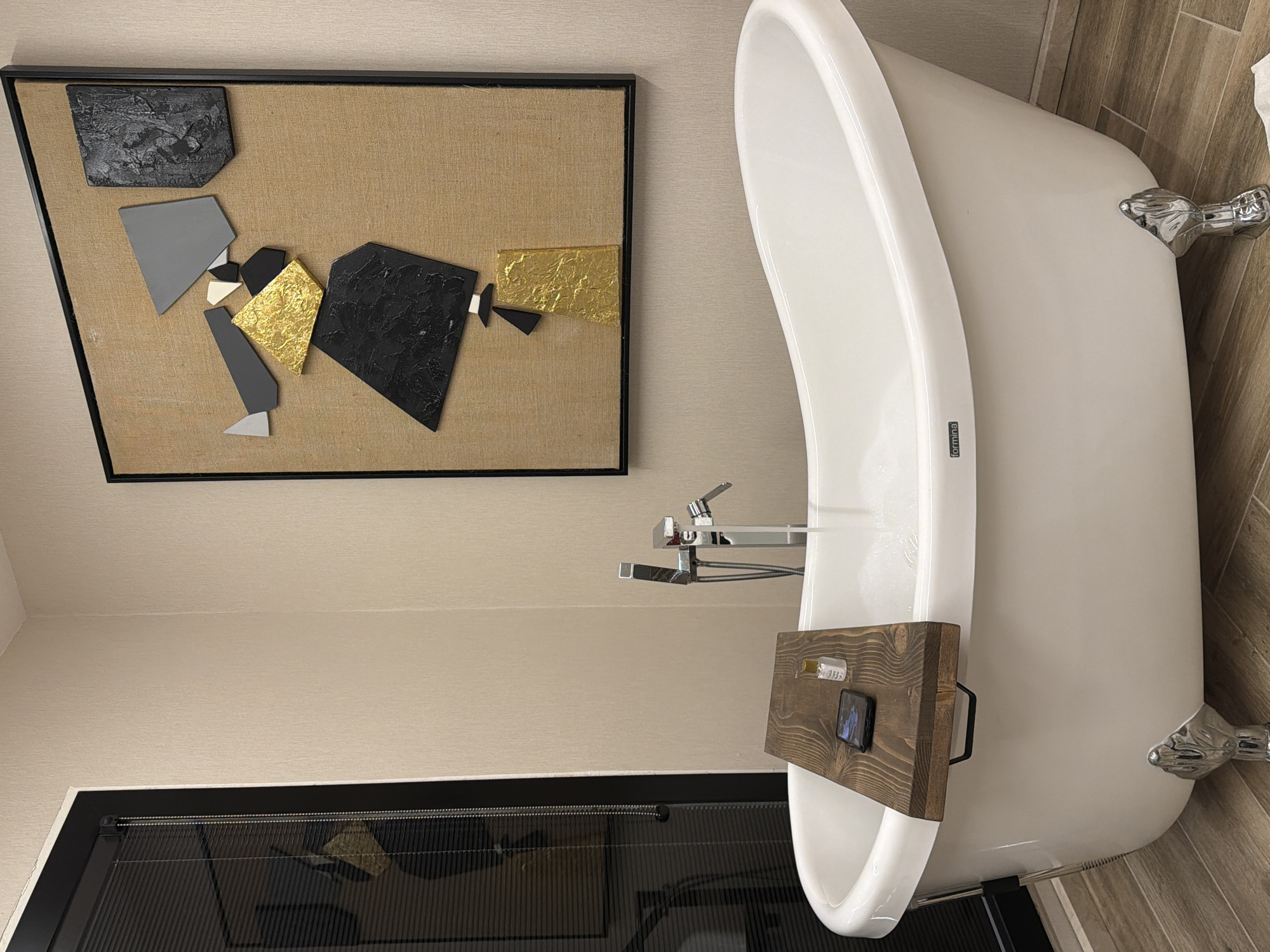 The image captures a modern bathroom interior, primarily featuring a freestanding white bathtub and an abstract artwork. The bathtub, positioned on the right side of the frame, is a deep, oval-shaped model with sleek chrome claw feet, two of which are visible. A dark wooden bath tray spans the tub's rim, holding a black smartphone with a lit screen and a small, clear bottle topped with a gold-colored cap. A tall, floor-mounted chrome faucet with a handheld shower attachment rises beside the tub. The brand name "formina" is visible on the side of the bathtub.
To the left, on a light beige, textured wall, hangs a large, framed abstract artwork. The artwork features an arrangement of various geometric shapes in black, gray, white, and reflective gold foil, set against a tan, woven canvas background within a simple black frame. The shapes are textured, with some black areas showing a raised pattern and the gold areas having a crinkled appearance.
The walls are uniformly light beige. The floor on the right side of the tub is covered with light brown wood-look tiles, arranged in a herringbone pattern. Below the tub, a dark, reflective surface, possibly a glass partition or a dark-tiled floor, shows a blurry reflection of the room. The scene depicts a serene and well-appointed space, with no specific activity taking place.