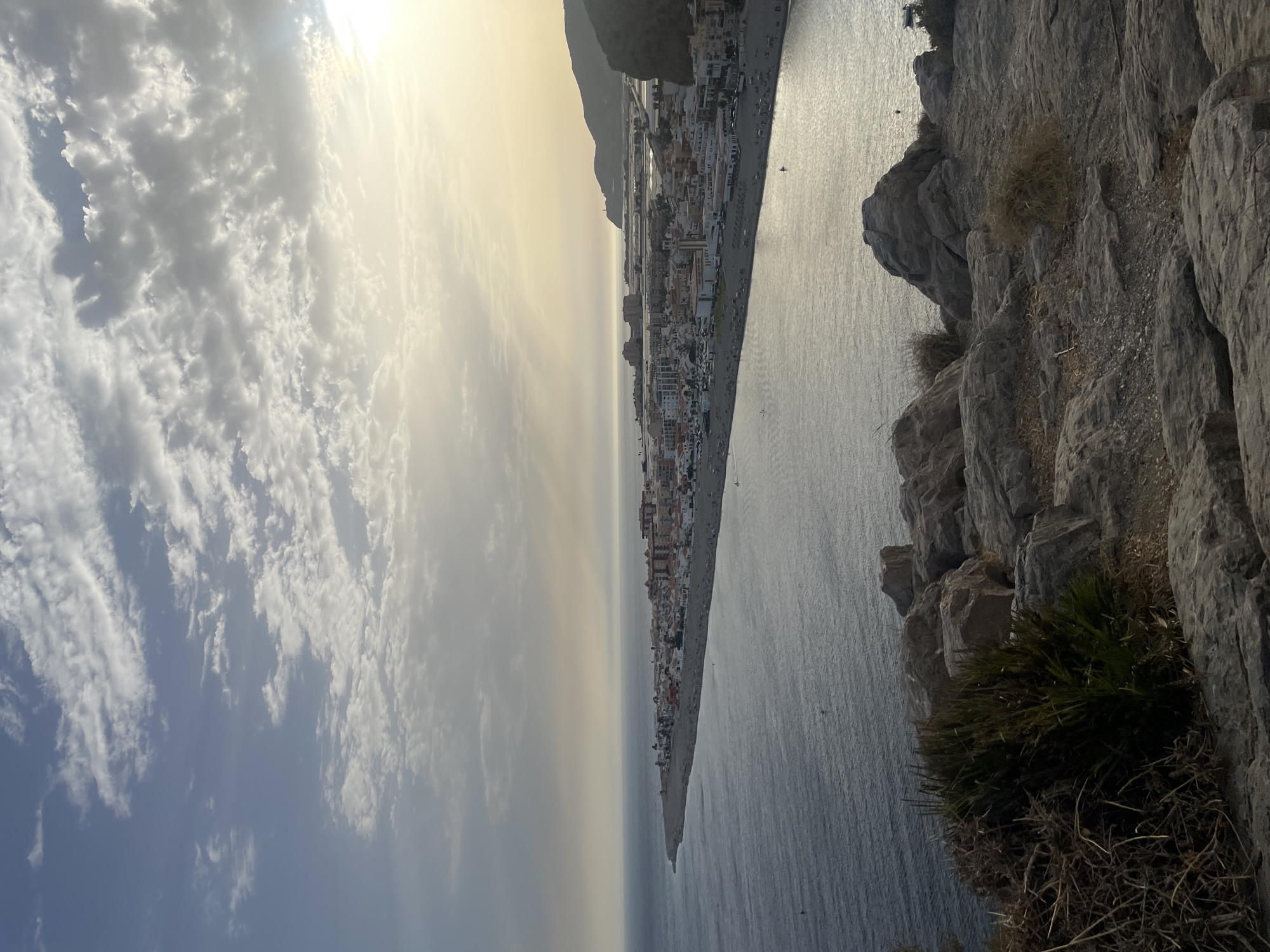 The image presents a coastal town, likely Gualchos, Spain, viewed from an elevated rocky vantage point. The foreground is dominated by rough, textured rocks with sparse dry vegetation and green shrubbery. Beyond this rocky outcrop, a wide, dark expanse of the sea stretches out, meeting a long, sandy beach lined with a densely packed town. Buildings in the town are predominantly white with some orange roofs, suggesting Mediterranean architecture. The beach is populated with numerous sunbathers and umbrellas. Further in the distance, hazy mountains form the backdrop to the town, with a lighthouse visible on the horizon. The sky is partly cloudy, with the sun breaking through, casting a warm, ethereal light over the scene, indicating either sunrise or sunset. The water is calm, reflecting the soft light.