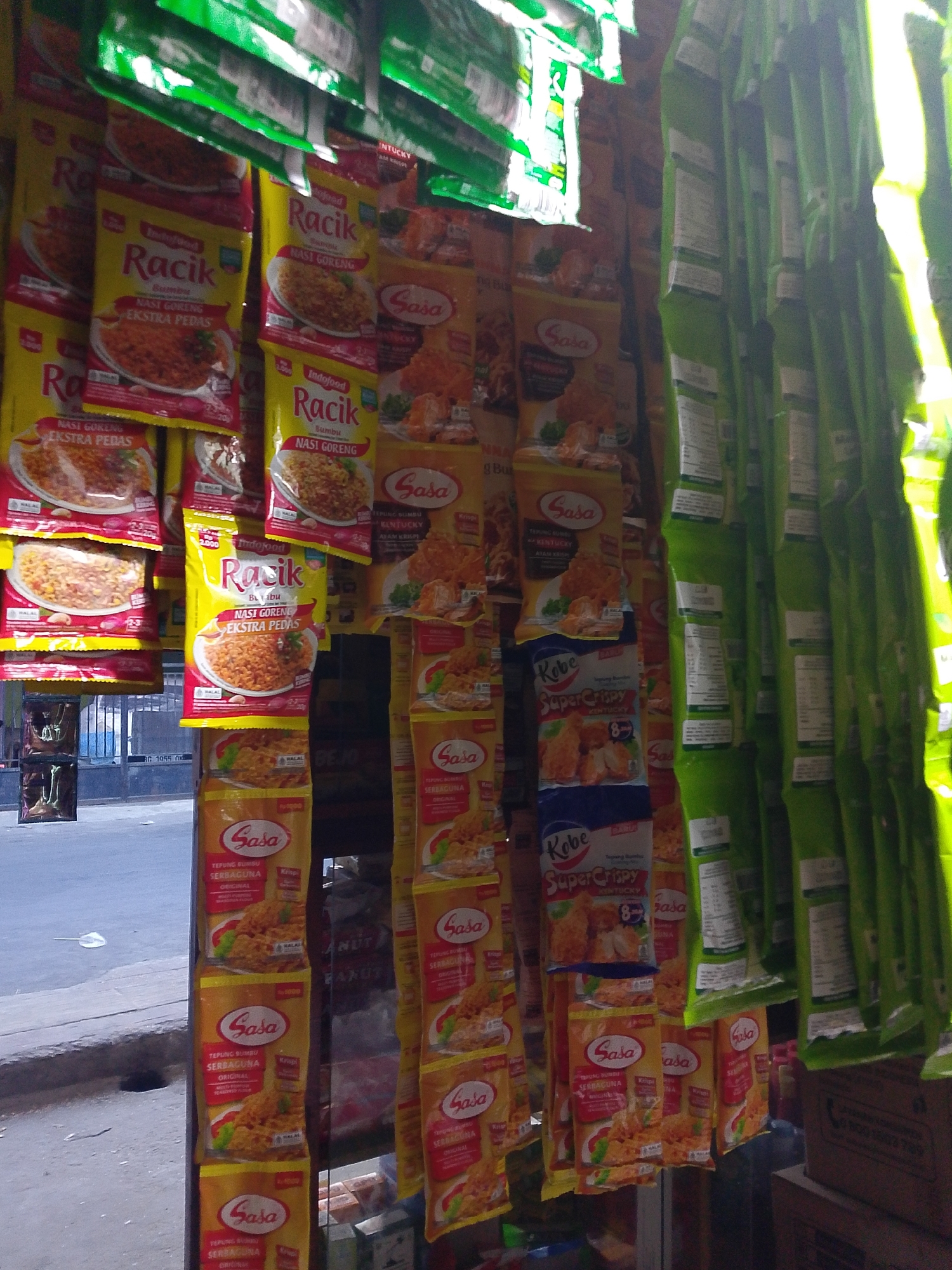The image shows a close-up of a stall selling various Indonesian food seasoning packets, likely in a market or small shop. The packets are brightly colored and feature images of food. Some of the brands visible include "Raik," "Sasa," and "Kobe Super Crispy." The stall appears to be outdoors, with a street and sidewalk visible in the background. The overall impression is of a busy, commercial environment typical of a Southeast Asian city.