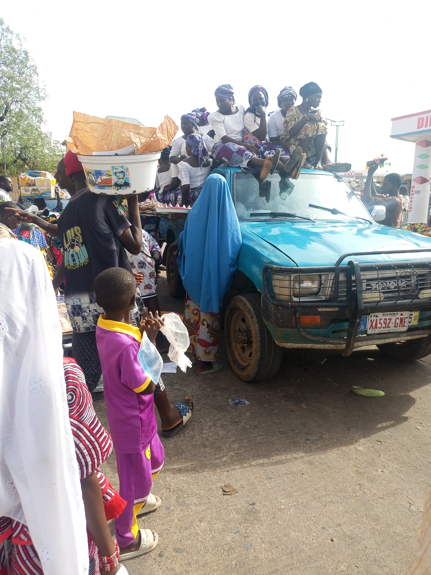 The image depicts a crowded street scene in Gombe, Nigeria. A light-blue Toyota pickup truck is the central focus, with several women sitting on its roof and hood. They are wearing various traditional Nigerian dresses and headwraps. A few individuals are gathered around the truck. A man carries a large white bucket on his head. A young boy in a purple outfit holds a plastic bag. A woman in a blue hijab stands next to the truck. A sign indicating "DI..." is visible in the background. The vehicle's license plate is XA592-GME. The overall impression is a festive, possibly celebratory, gathering.