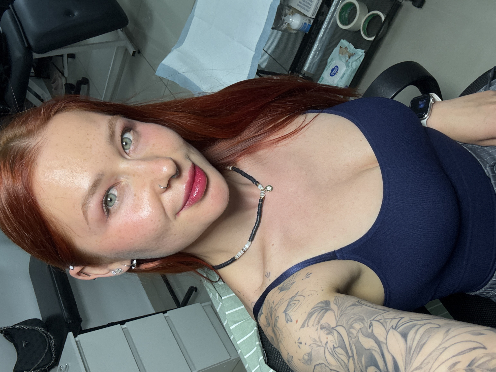 A woman with red hair, fair skin, and freckles is photographed indoors. She has green eyes, a nose ring, and multiple earrings. She wears a dark blue top and a black beaded necklace. A tattoo is visible on her left arm. The background suggests a medical or cosmetic setting, with medical supplies and equipment visible. The photo may have been taken in Rostov-on-Don, Russia.