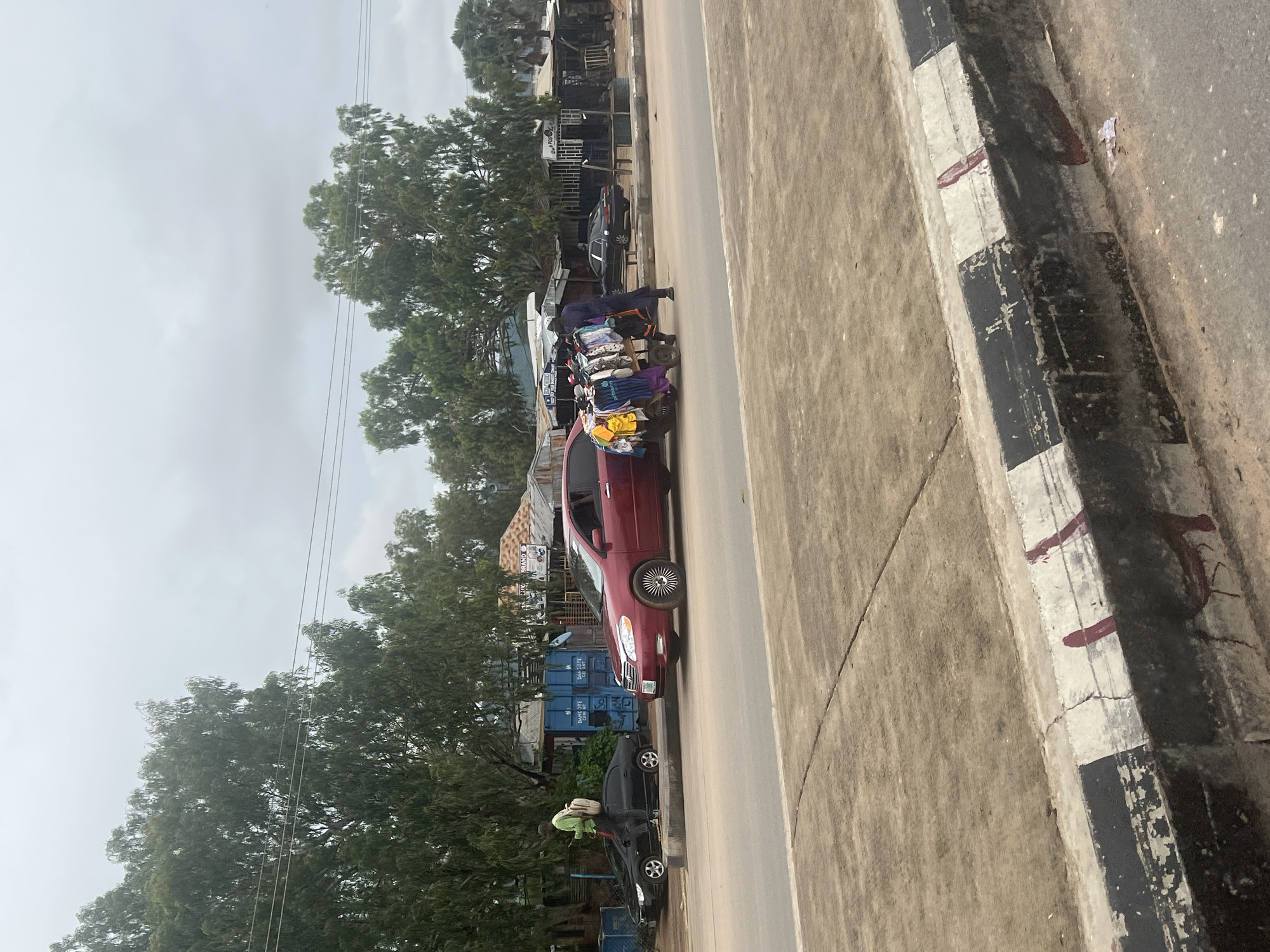 This is a street scene in Jalingo, Nigeria, under a cloudy sky. In the foreground, a road with a concrete median strip painted with black and white stripes runs horizontally. Beyond the median, a red sedan is parked, its side visible, with a cart loaded with clothing beside it. A person in dark clothing stands near the cart, appearing to be attending to the merchandise. To the left of the red car, a black sedan is parked. Behind the vehicles, there are various structures, including what appear to be market stalls and shipping containers, shaded by trees. A person in a light green shirt and a backpack is walking away from the black sedan. Overhead, power lines are visible against the overcast sky. The overall scene depicts a commercial roadside area.