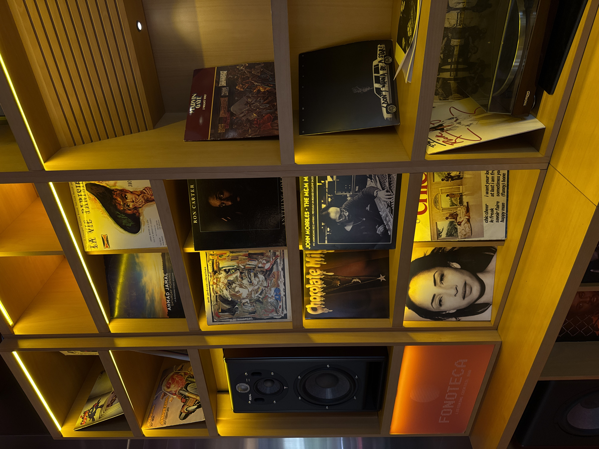 This is a medium shot of a wooden shelf unit displaying vinyl records and a speaker. The shelf unit is illuminated by warm yellow LED lights. On the far left, a black speaker sits on a shelf. Above it are several vinyl records, including one with a comic book-style cover featuring a superhero. To the right, more vinyl records are displayed, with album art varying from portraits of musicians to abstract designs. One record has the title "La Vie Parisienne" on its cover. Another features a portrait of a woman. Towards the bottom right, a record player with the brand name "Thorens" is visible. A sign with the text "FONOTECA LISTENING COCKTAIL BAR" is also visible on the bottom left shelf. The overall impression is a well-curated display of music and audio equipment in a bar setting. The location is stated to be Yerevan, Armenia.