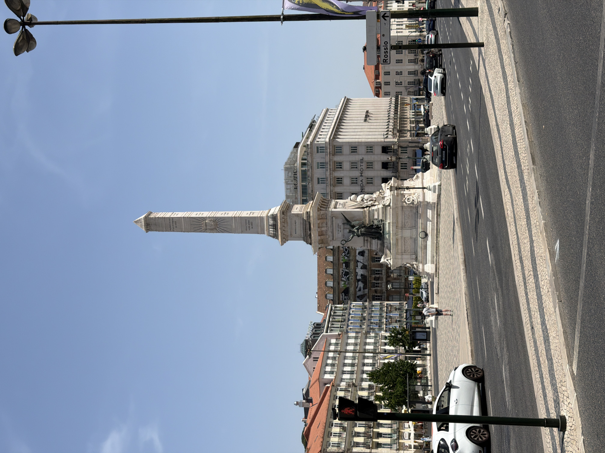 The image shows Rossio Square in Lisbon, Portugal on a clear day. In the foreground, a street with traffic and pedestrians is visible. A white car is parked on the left, and several other cars are driving on the road. A pedestrian crossing with a traffic light for pedestrians is also present. In the background, the Rossio Monument, an obelisk, stands prominently in the center of the square. Several buildings surround the square, including the Rossio Hotel. A lamppost with multiple lights is on the right side of the image, and a street sign points to "Rossio." The architecture is typical of Lisbon, with colorful buildings and intricate facades.