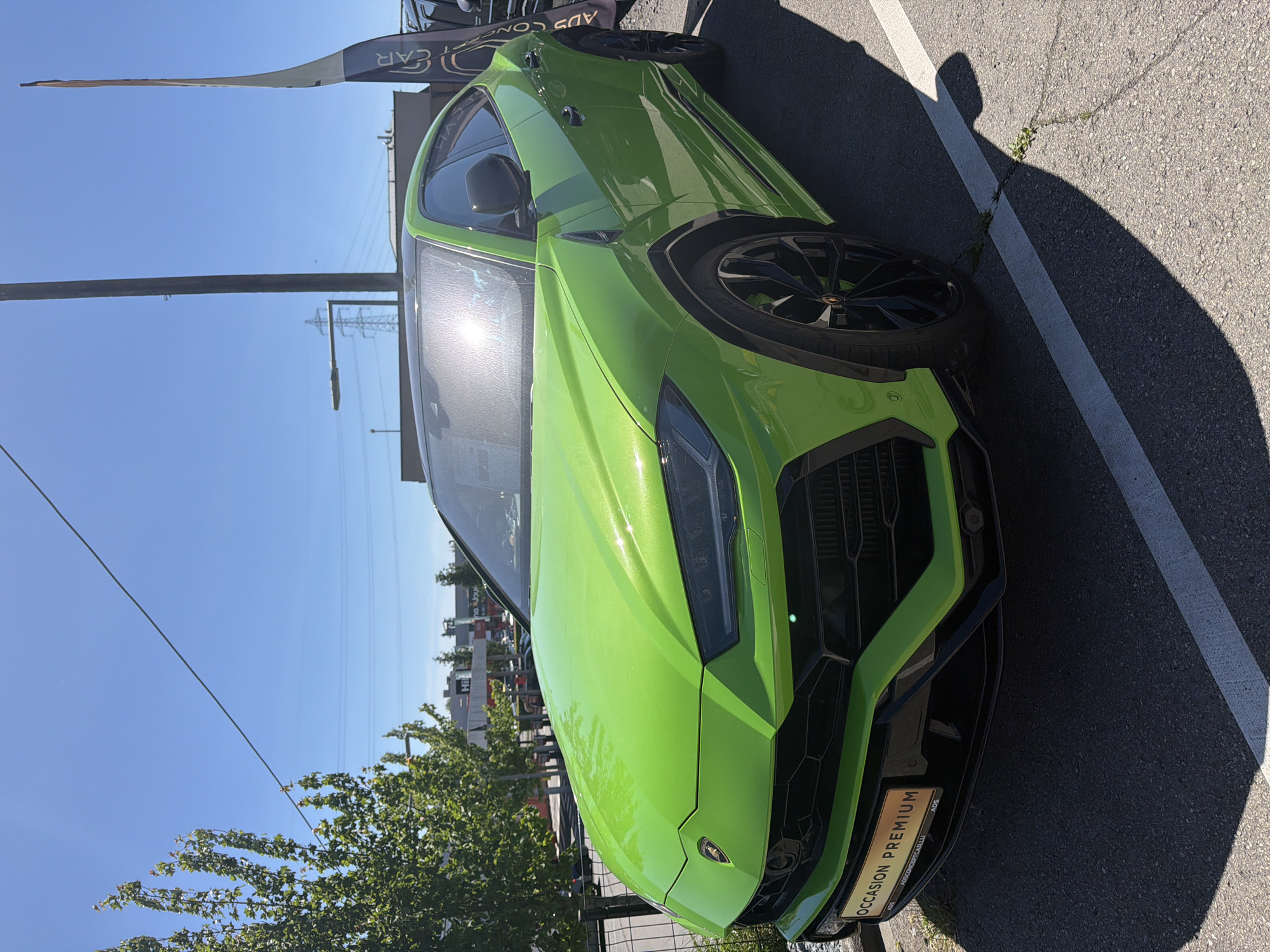 A bright green Lamborghini Urus is parked in an outdoor lot in Mondercange, Luxembourg. The vehicle is a modern SUV with a sleek, aerodynamic design and black rims. The car has a panoramic sunroof, and the front of the car is illuminated by the sun, creating a bright reflection on the windshield. A license plate reading "OCCASION PREMIUM" is attached to the front bumper. The background shows trees, buildings, and a clear blue sky, suggesting a sunny day. A tall, thin flag with the text "ADS CONCEPT CAR LU" is visible to the right of the vehicle, indicating the location is likely a car dealership. The ground is paved with asphalt, and a white line marking a parking spot is visible in the foreground.