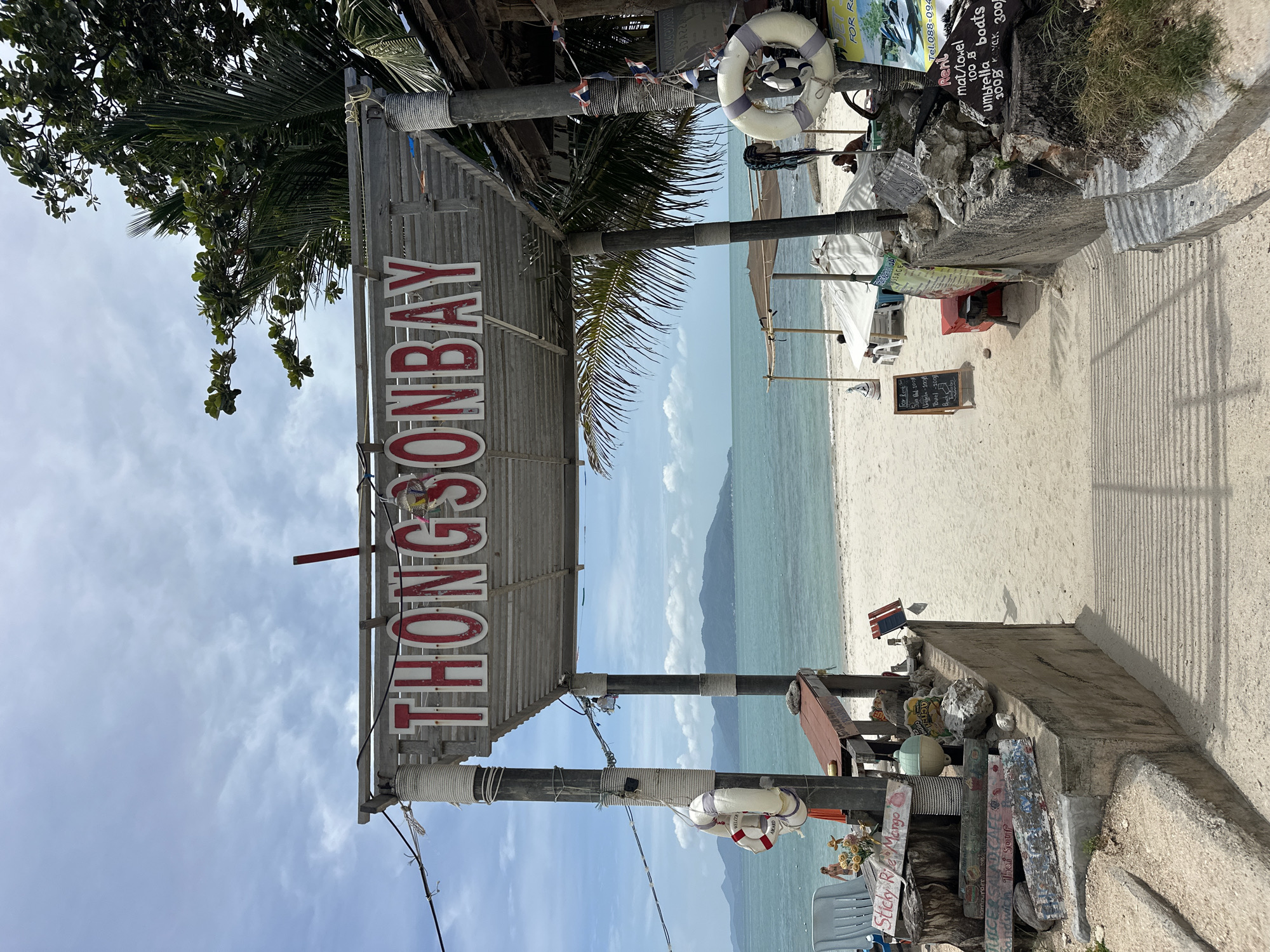 A sign that reads "THONGSONBAY" is displayed prominently in front of a beach. The sign is made of red lettering on a white background, and it is mounted on a wooden structure with metal supports. The beach itself is a white sandy expanse, with turquoise waters stretching out to the horizon. In the background, a chain of islands can be seen. Several beach umbrellas and chairs are set up on the sand, suggesting a resort or tourist area. There are also a few people visible, some of whom are in the water. The overall impression is one of a tropical paradise, perfect for a relaxing vacation.