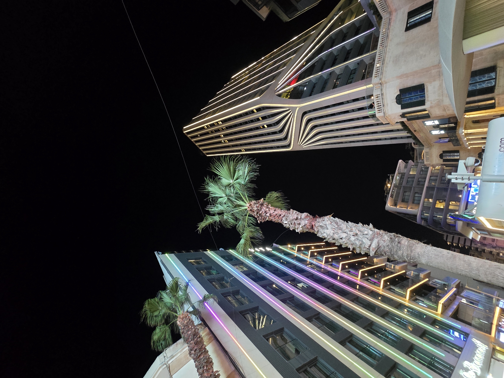 The image captures a nighttime view of a street scene in Saint Julian's, Malta, featuring modern architecture illuminated by colorful lights and a stark contrast with the dark night sky. The foreground is dominated by two palm trees with rough, textured trunks and green fronds, one leaning towards the left and another towards the right of the frame.
To the left, a tall building with a dark facade is adorned with vertical stripes of vibrant LED lights in shades of purple, pink, green, and yellow, creating a striking visual effect. The building has multiple floors with visible windows.
To the right, a more distinctively shaped modern skyscraper rises, its facade outlined with numerous thin, bright yellow lights that follow a patterned, curved design, converging towards the top. The base of this skyscraper appears to be a more traditional, light-colored stone building with windows and balconies, featuring architectural elements like columns and archways.
In the background, other buildings of varying heights and architectural styles are visible, some with illuminated windows, adding depth to the urban landscape. The overall impression is one of a lively, contemporary city at night, with an emphasis on architectural design and artificial lighting. The scene is devoid of visible people or any discernible human activity.