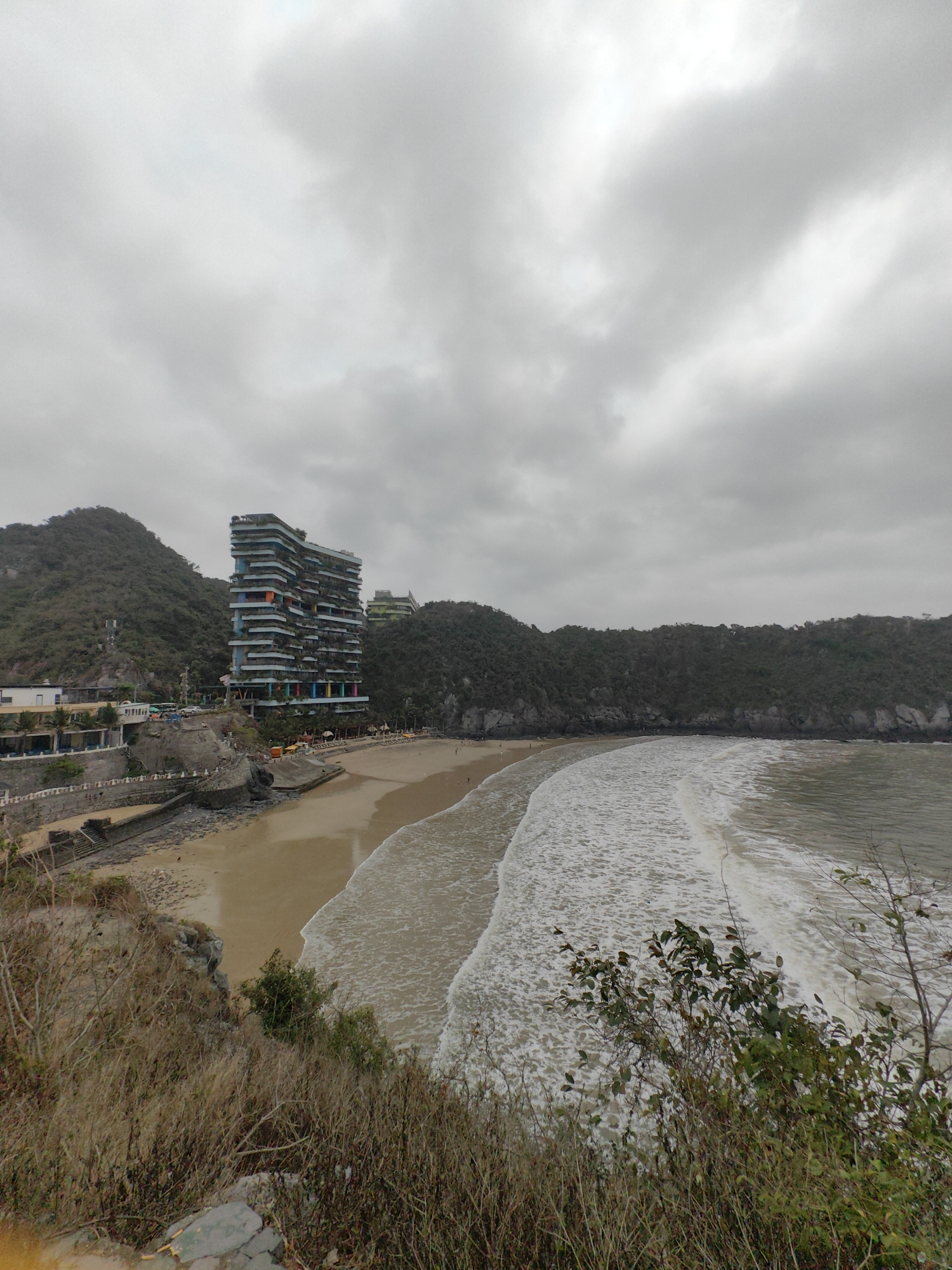 Captured from an elevated viewpoint, the image presents a coastal scene in Hải Phòng, Vietnam under an overcast sky. Dominating the view is a sandy beach curving along the bay, with waves breaking onto the shore. A notable modern architectural building with colorful accents stands near the beach, adjacent to a smaller structure. The bay is framed by verdant, hilly terrain on either side. The foreground contains dry vegetation on the slope leading down to the beach. People are sparsely populated along the beach.