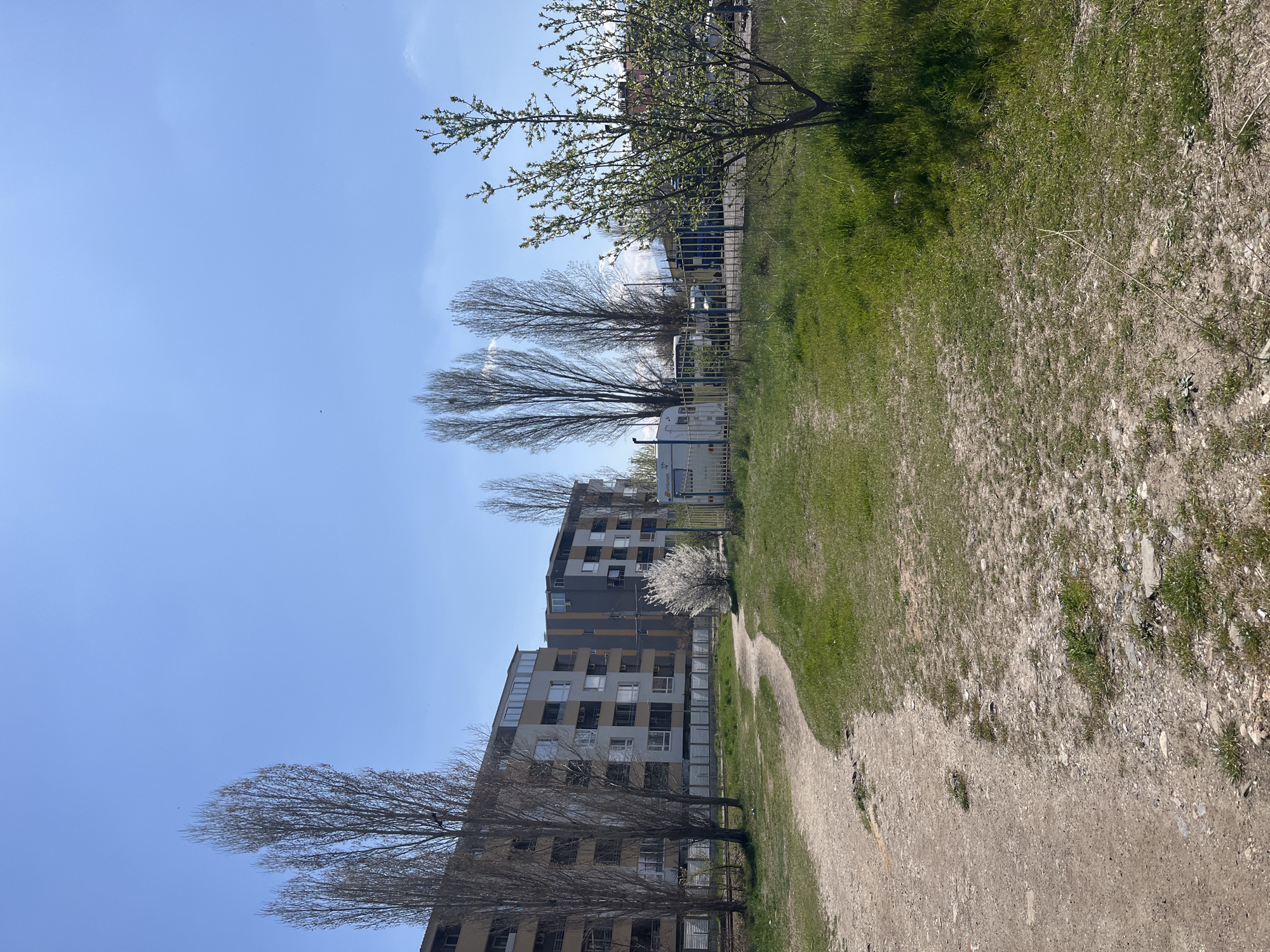 The image depicts a view in Sofia, Bulgaria, featuring a multi-story apartment building, trees, a grassy area, and a dirt path under a blue sky. The building is constructed of a light-colored material with rows of windows. Trees, some bare and others budding, are positioned along the building and the path. A section of green grass transitions to a worn dirt path. A white vehicle with a blue top is parked near the building. A chain-link fence runs parallel to the path and building.