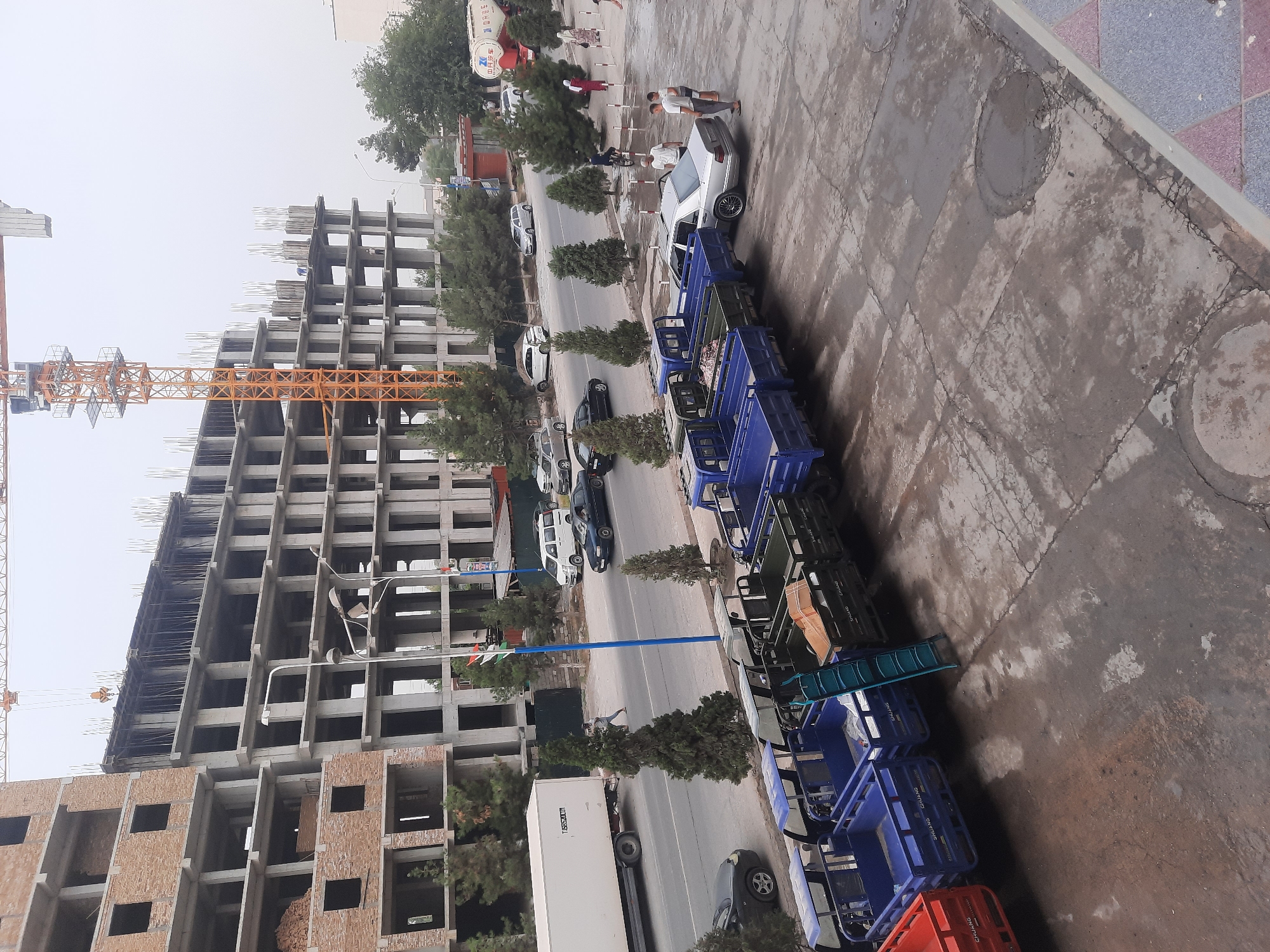 The image captures a street scene in Hulbuk, Tajikistan, with a focus on a construction site and a row of parked utility vehicles. In the background, a multi-story building is under construction, with visible concrete framework and scaffolding. A large yellow construction crane dominates the skyline. The street is lined with trees and appears to be an urban area.
In the foreground, a line of blue three-wheeled utility vehicles, commonly known as "tuk-tuks" or auto-rickshaws, are parked along the sidewalk. Some of these vehicles have their cargo beds filled with various items, including what appears to be fabric or tarp. One red utility vehicle is also visible at the far left. A silver sedan is parked next to some of the utility vehicles. Several individuals are present in the scene, including some standing near the silver car and others walking or standing on the sidewalk. A white delivery truck is parked on the street to the left of the construction site. The ground surface is a mix of concrete and asphalt, with some patches appearing wet. Flags are visible on poles along the street. The sky is overcast.